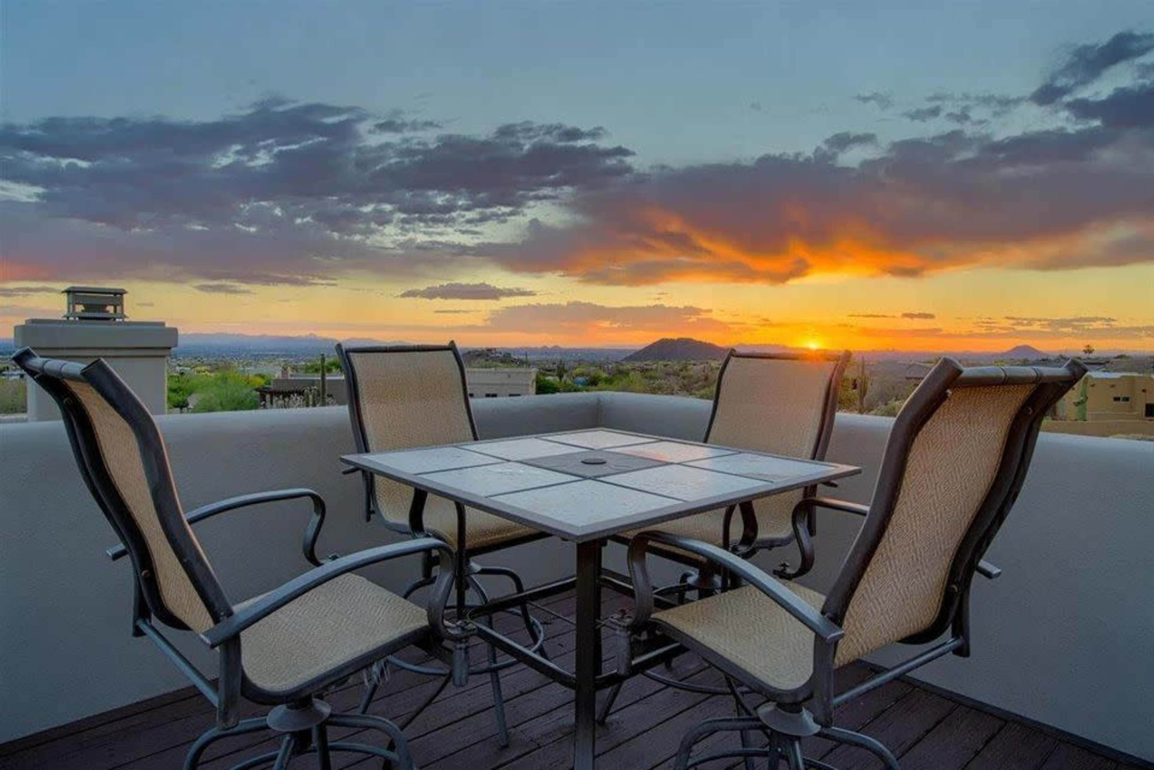 A patio set with four chairs surrounds a square table, overlooking a sunset with mountains and clouds in the background.