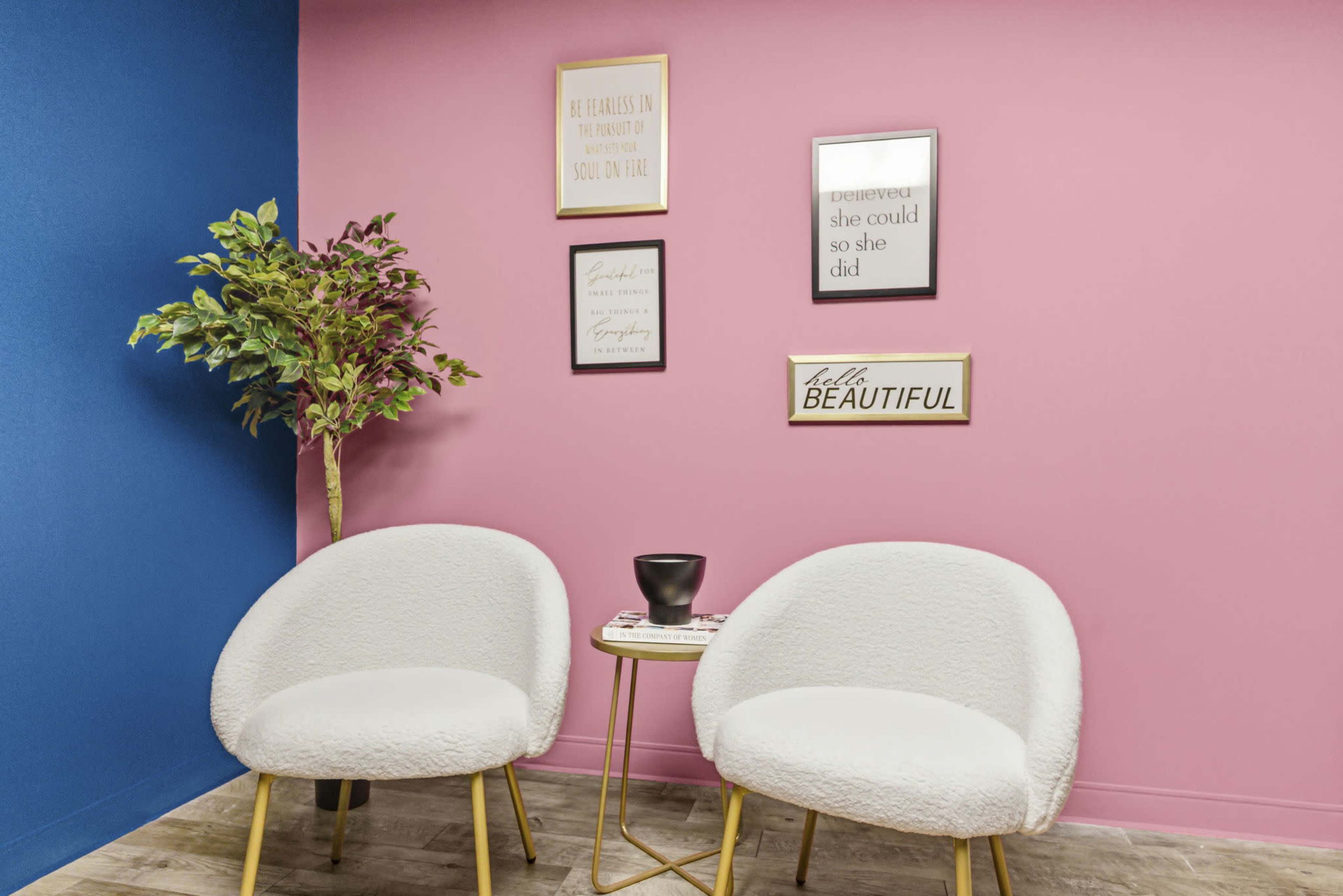 A cozy seating area features two white chairs and a small gold side table against a backdrop of pink and blue walls, adorned with framed inspirational quotes.