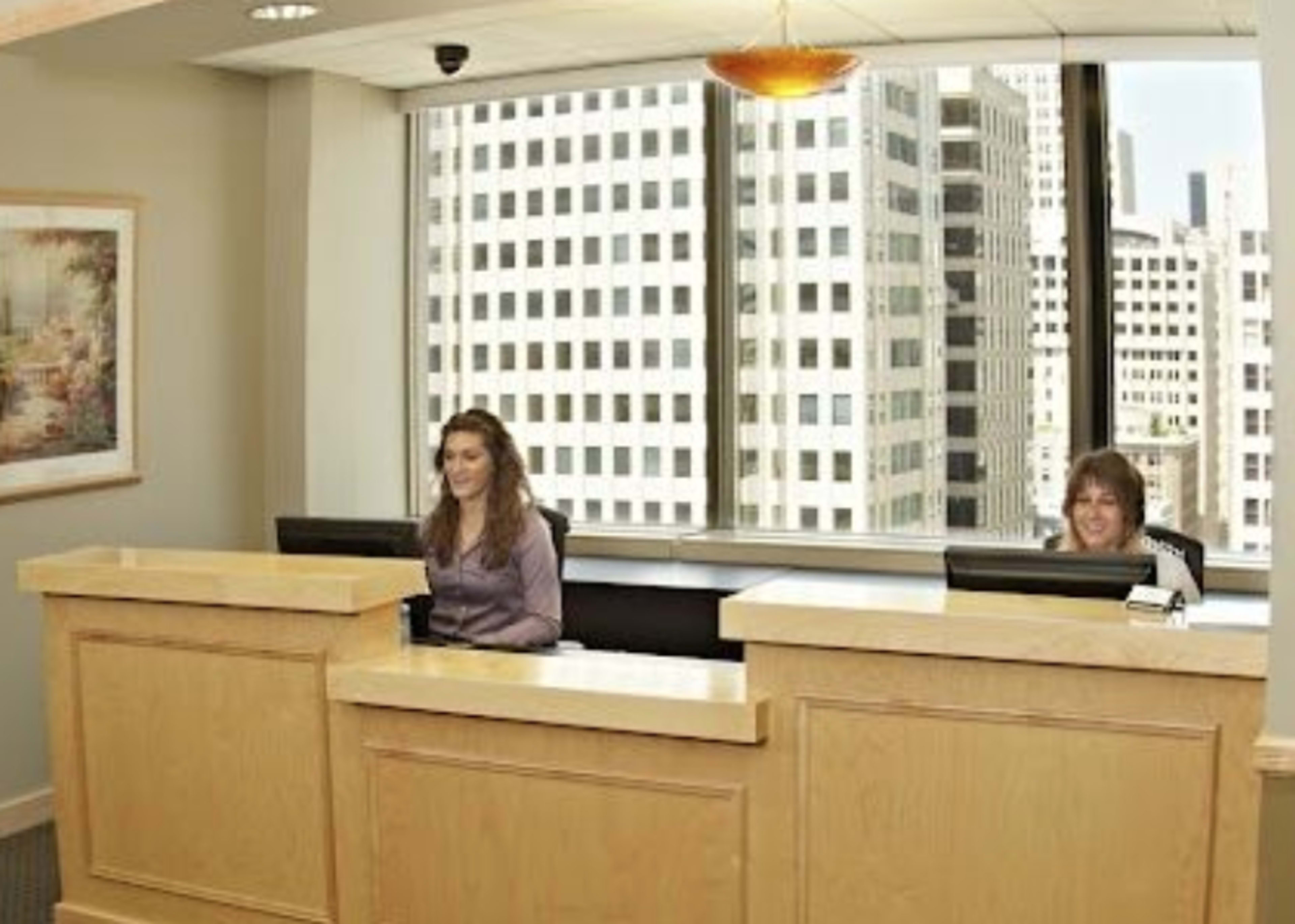 Two receptionists sit behind a wooden front desk in an office with large windows overlooking city skyscrapers.