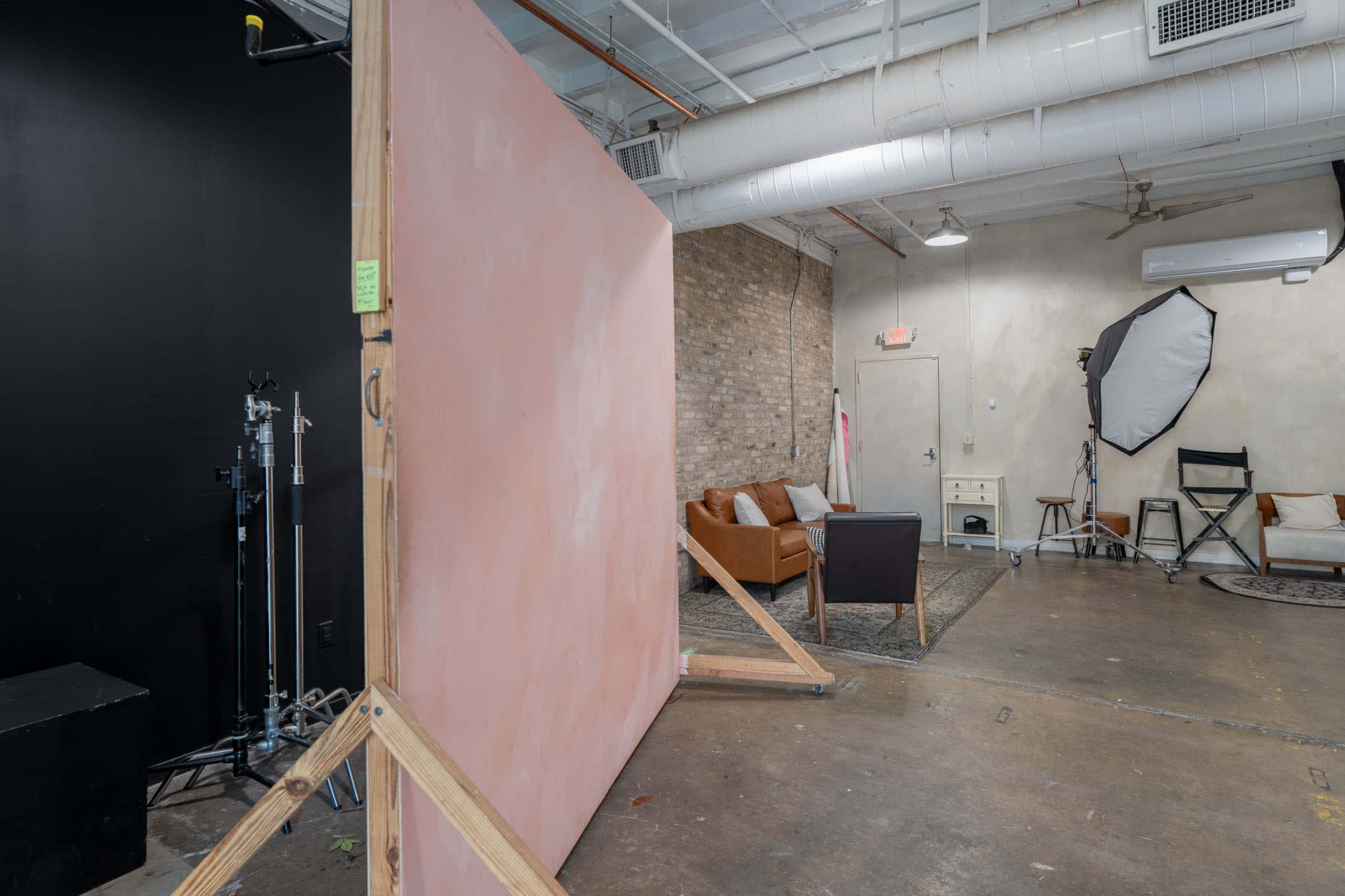 The image shows a photography studio with a large pink backdrop, a brown leather couch, and various equipment including a light source and a director's chair.