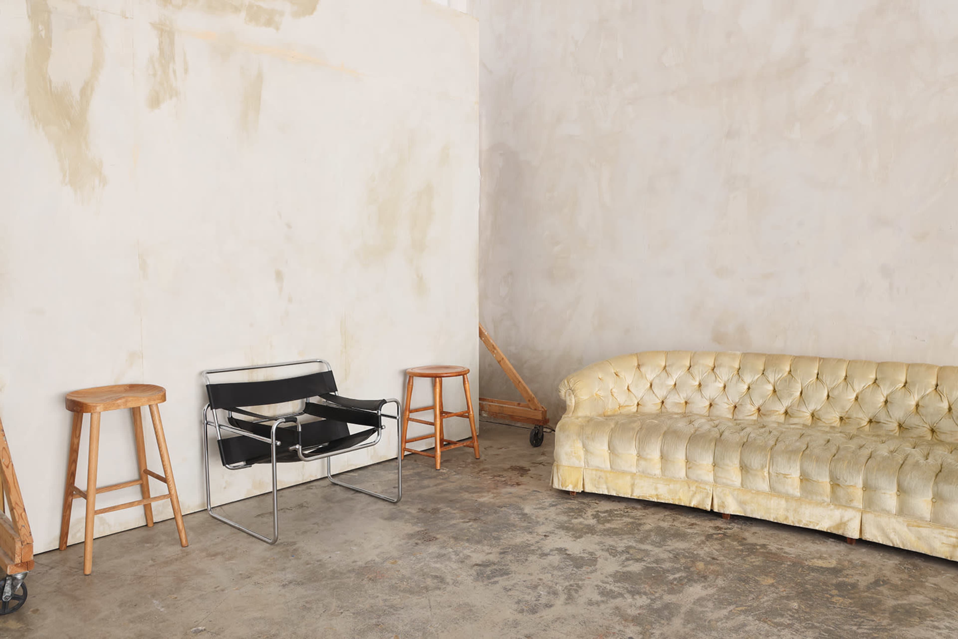 The image shows a minimalist interior with a beige wall, a worn yellow tufted sofa on one side, a black chair in the center, and two wooden stools positioned nearby.