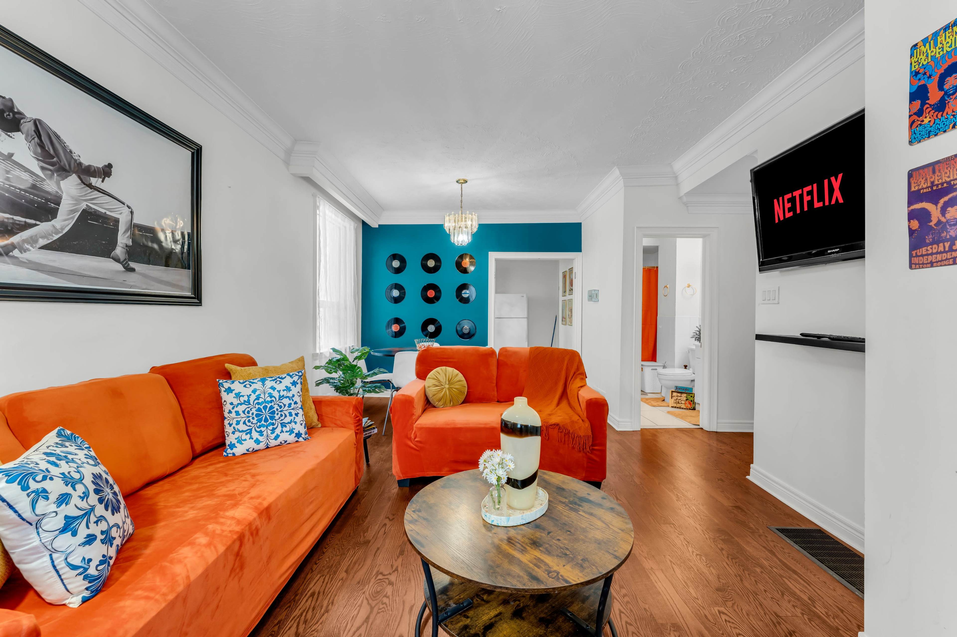 The living room features two orange sofas, a round coffee table, and a television mounted on the wall displaying the Netflix logo, with vinyl records arranged on the blue wall.