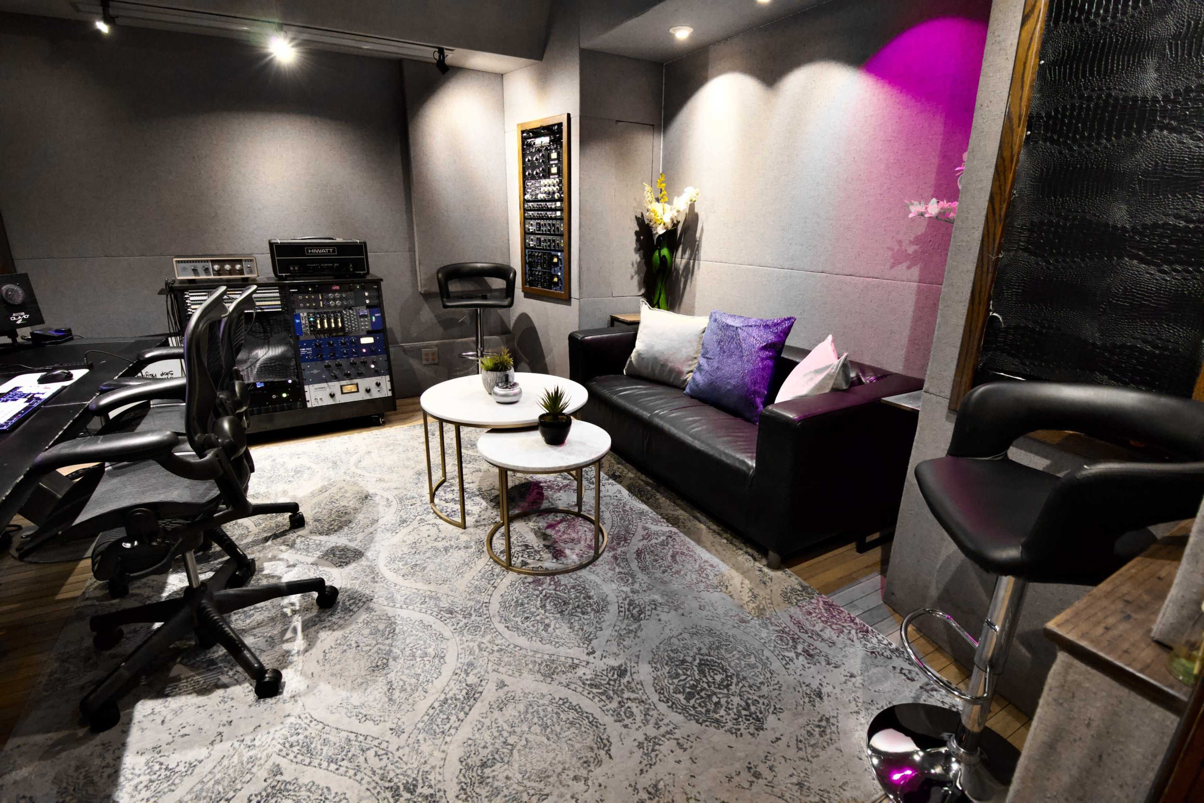 A modern recording studio featuring a black couch, two circular coffee tables, and audio equipment against a wall with gray soundproofing panels.