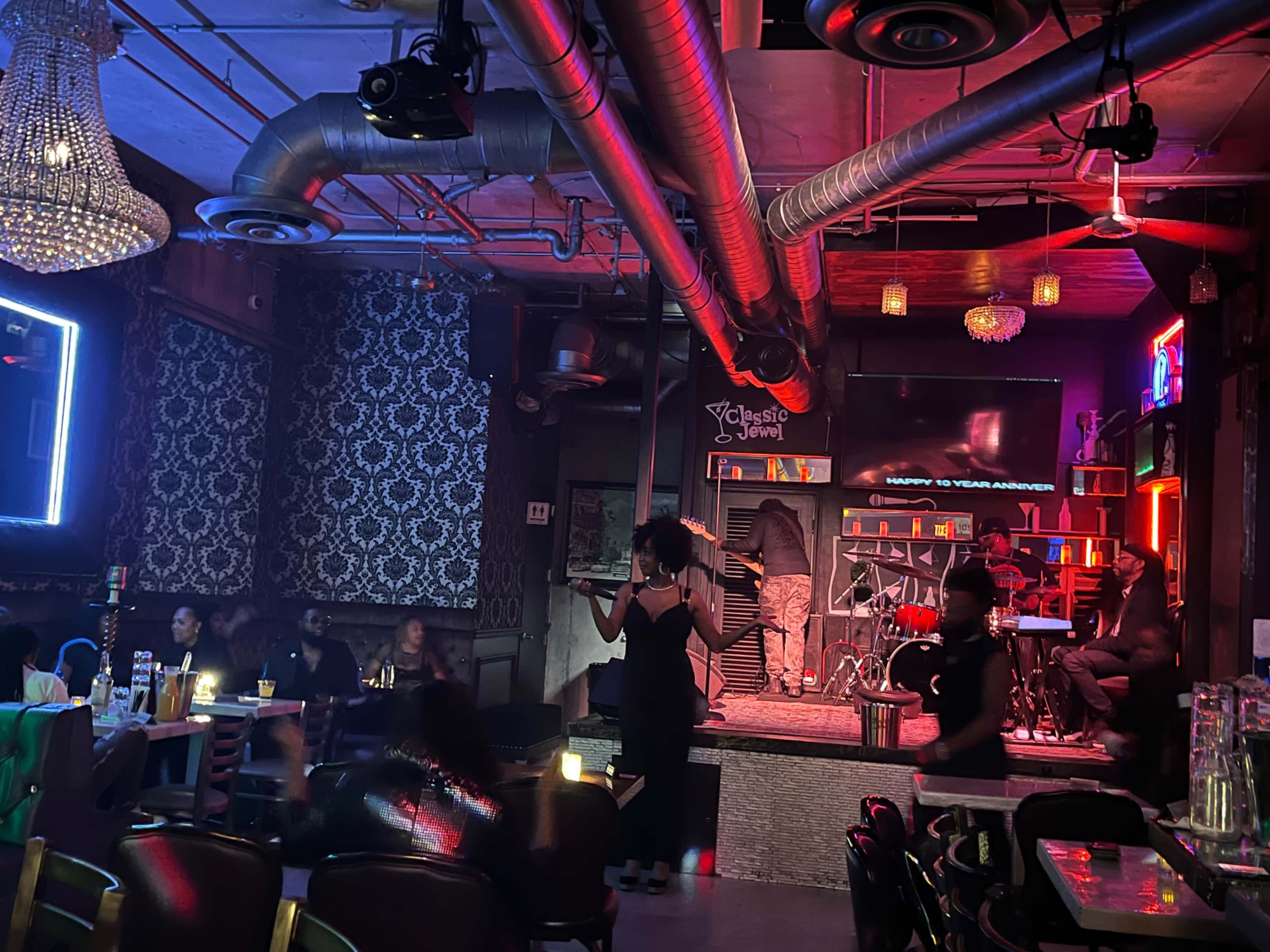 A singer performs on stage in a dimly lit bar while an audience watches and musicians play in the background.