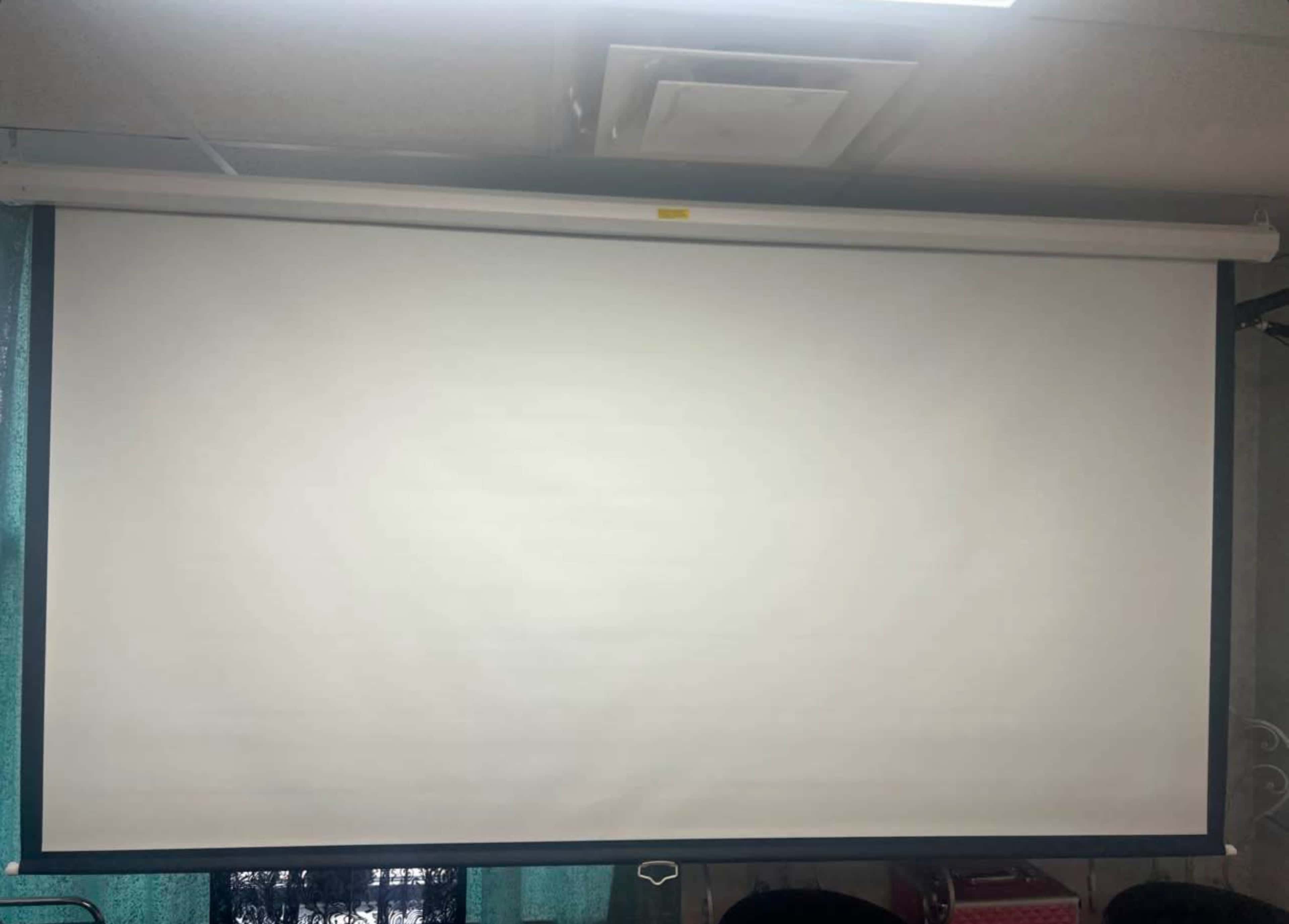 A white projection screen is mounted on a wall in a room with teal curtains in the background.