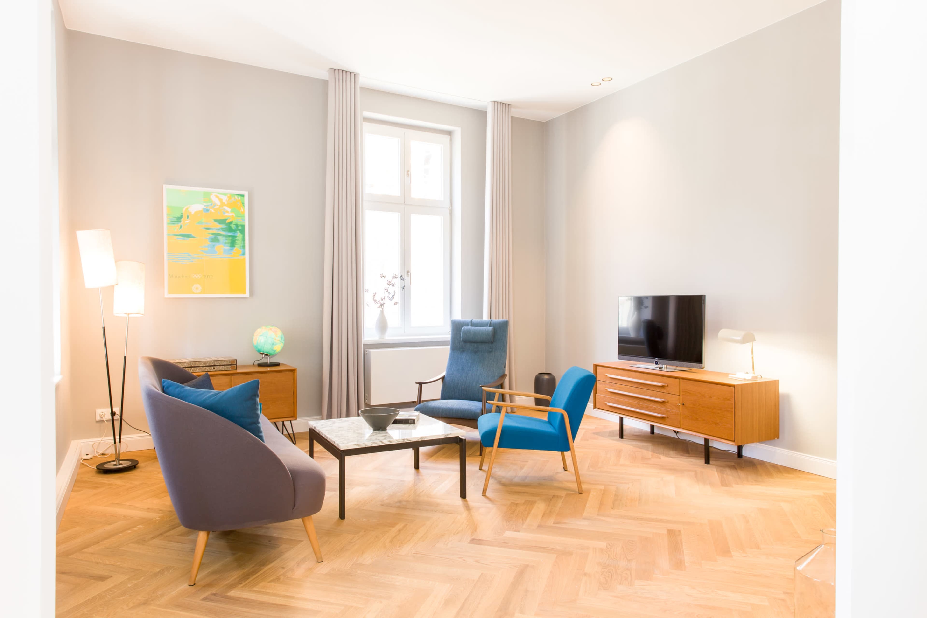 A modern living room features two blue armchairs, a wooden coffee table, a television on a console, and a large window with light gray walls and hardwood flooring.
