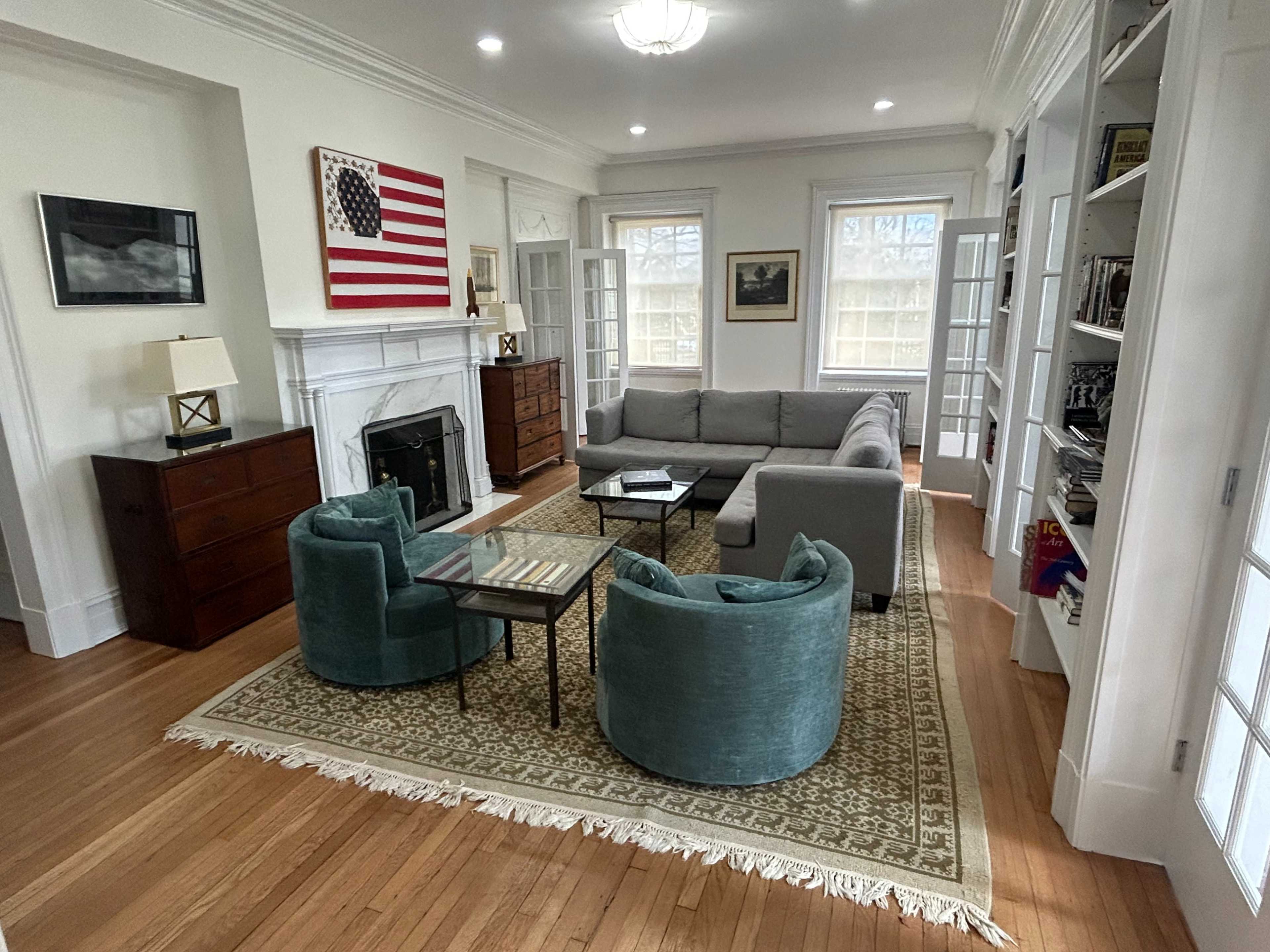 The image shows a living room with a gray sectional sofa, two teal chairs, a glass coffee table, and a large area rug, complemented by an American flag artwork on the wall.