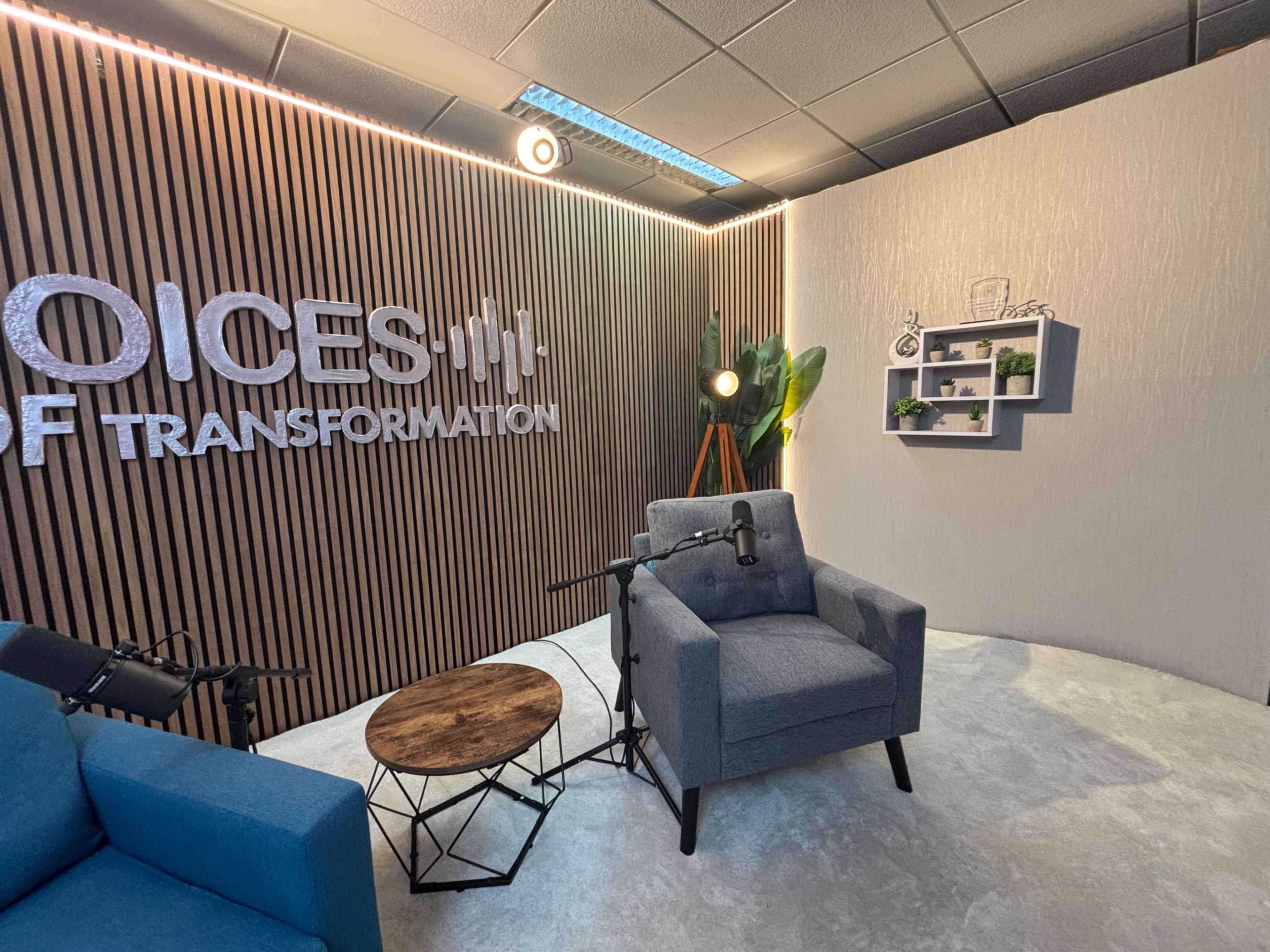 The image shows a modern studio setup featuring a gray armchair, a wooden coffee table, microphones, and a backdrop with the text "Voices of Transformation" against a wooden wall.