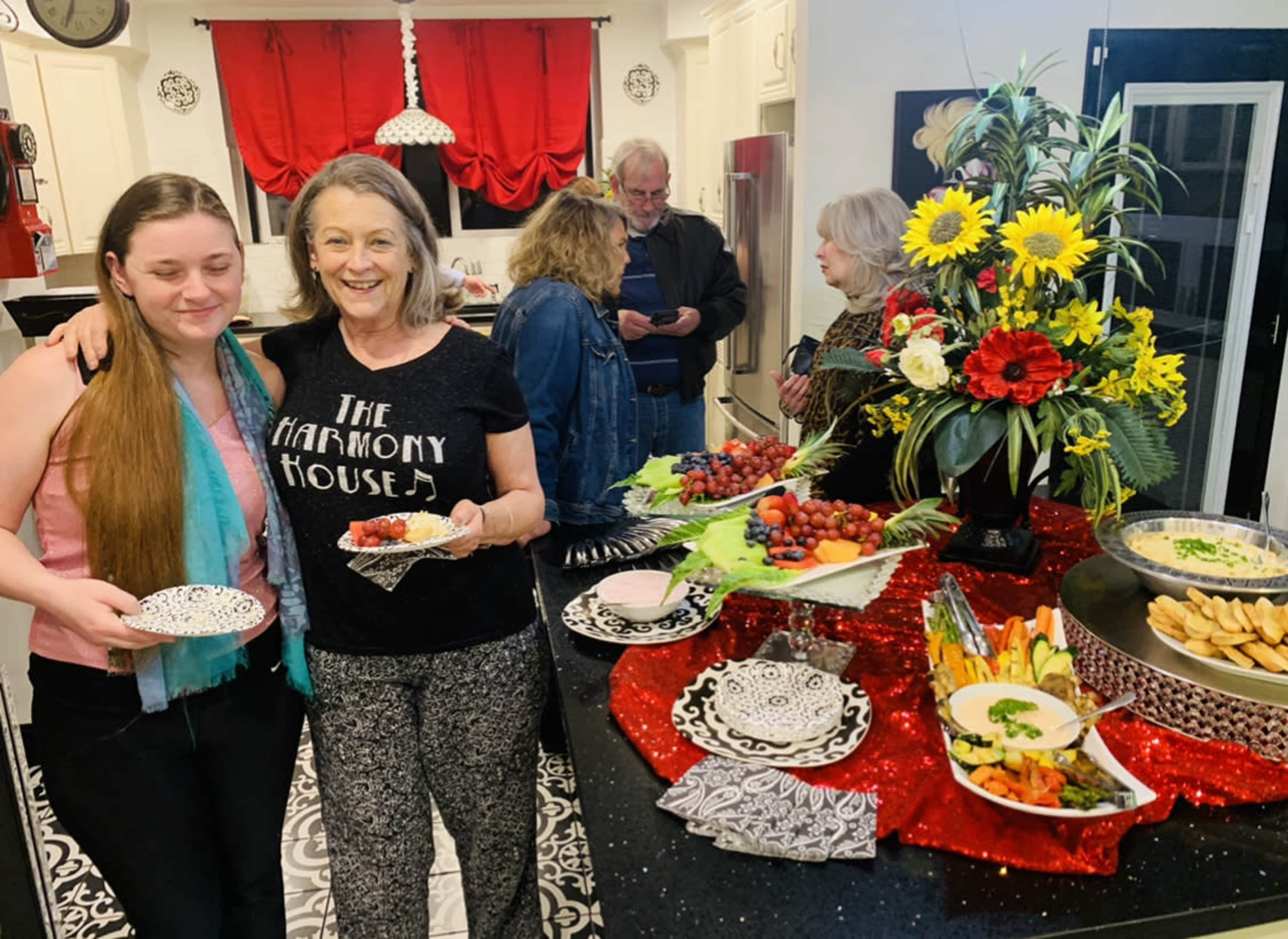 A group of people gathers in a kitchen with a decorated table featuring various appetizers and a floral arrangement.