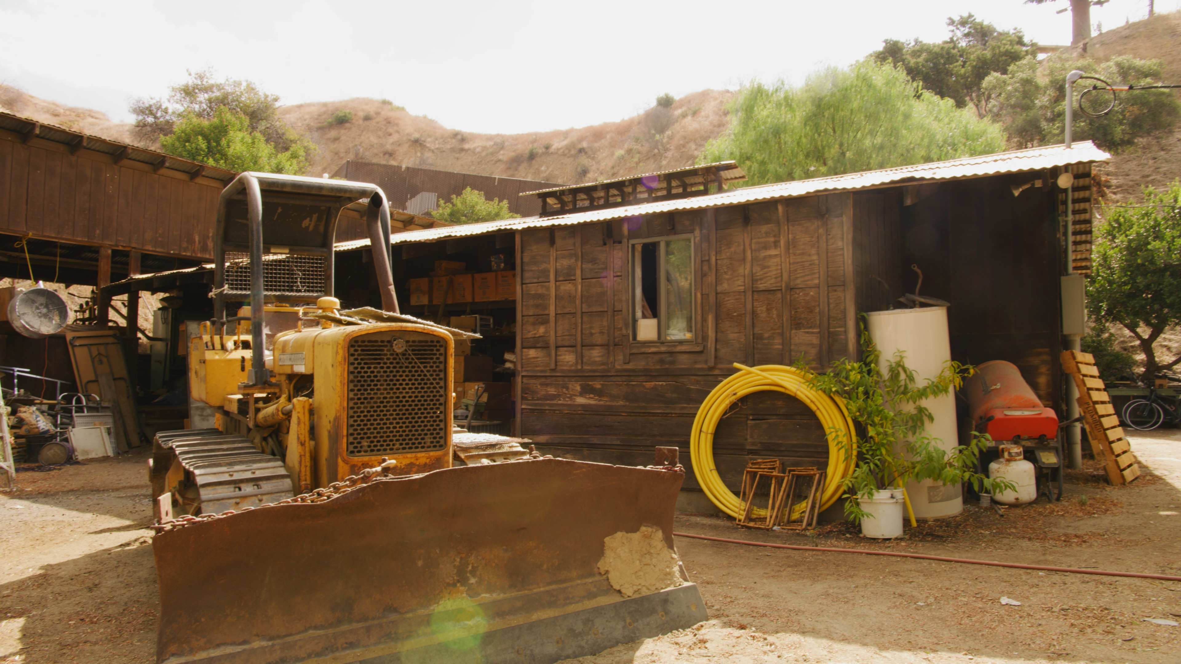 A yellow bulldozer is parked in front of a wooden shed with a mountain backdrop and several items scattered around the yard.