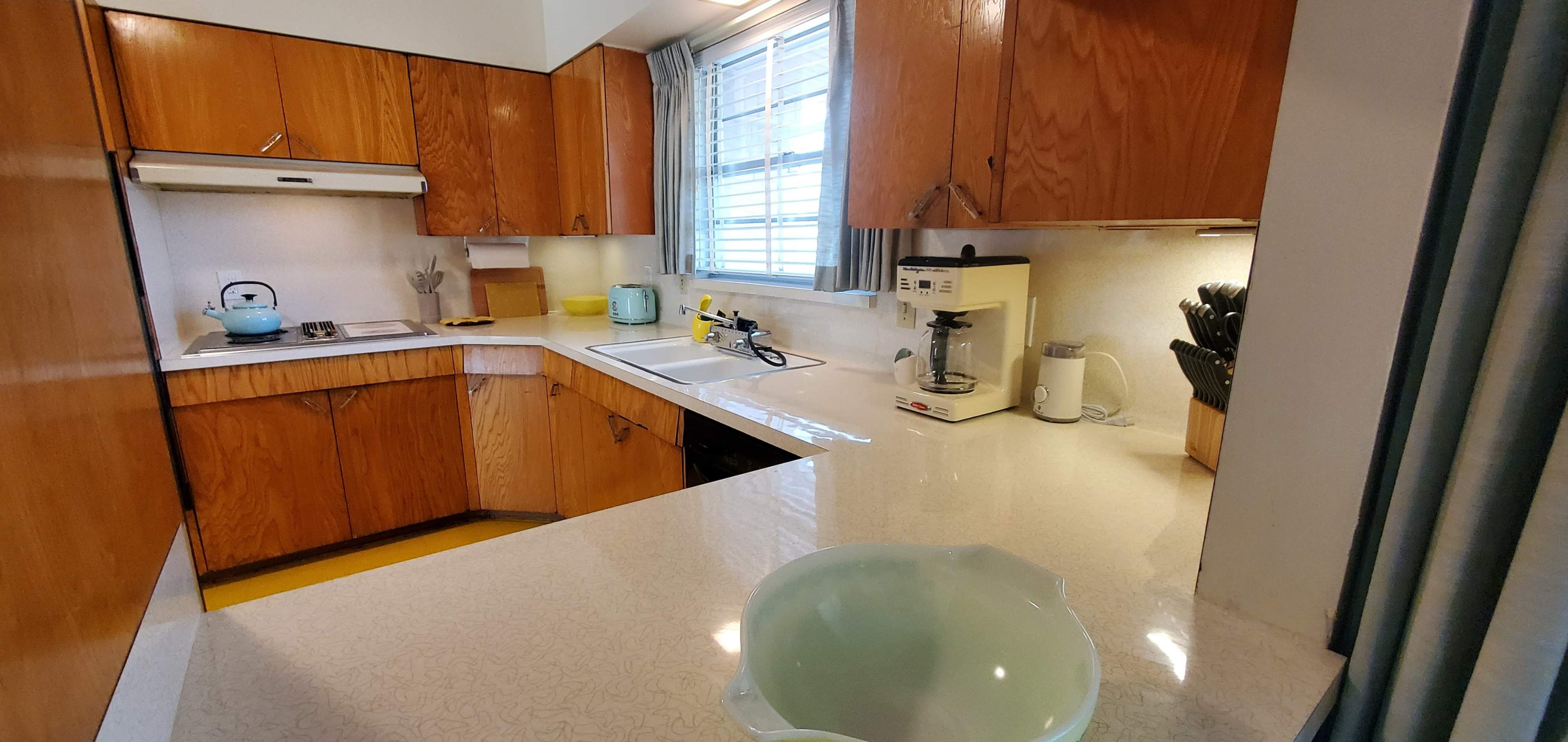 A kitchen featuring wooden cabinetry, a window with light blue curtains, and various kitchen appliances on a countertop.