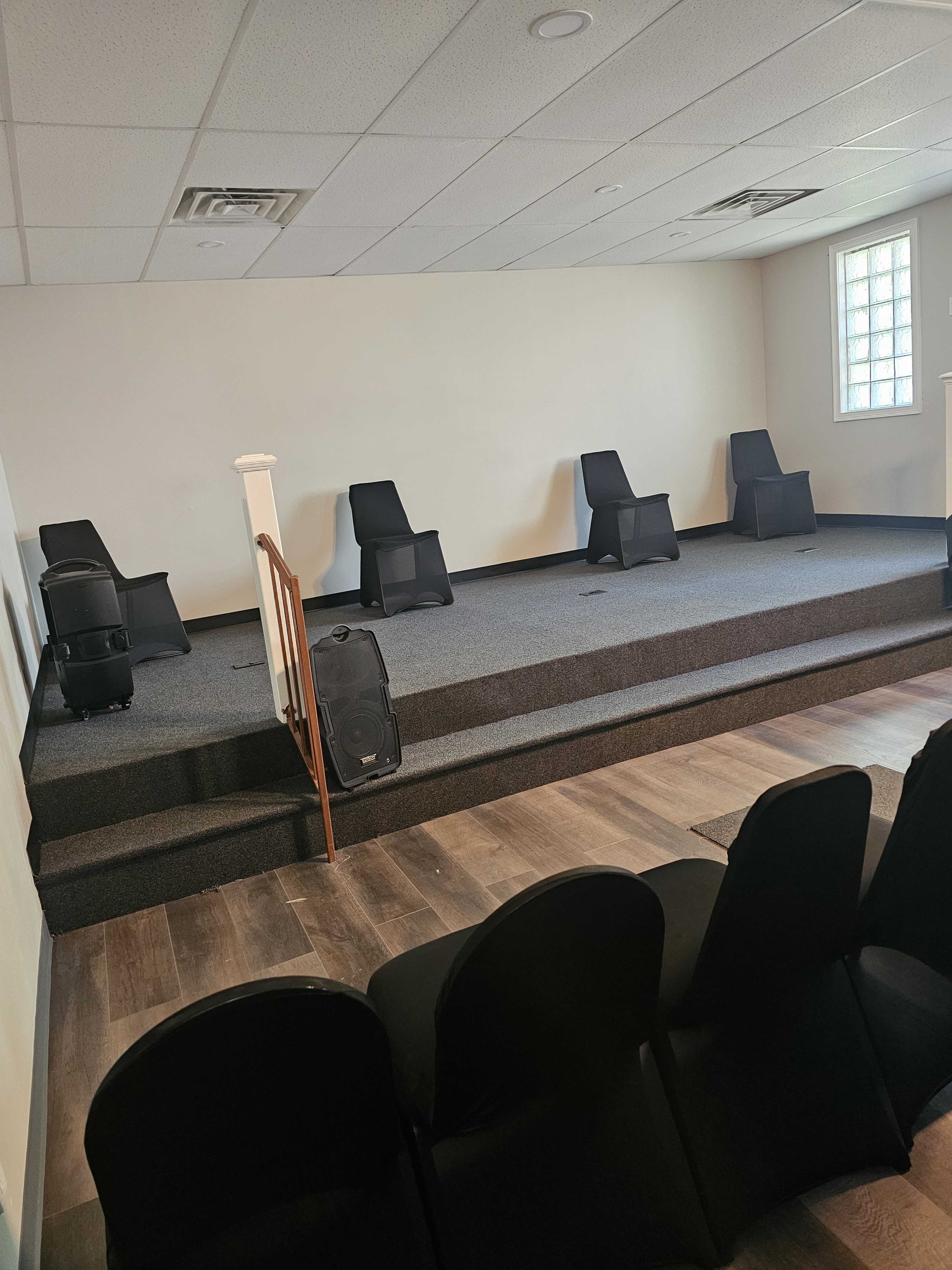 The image shows a small stage with five black chairs placed on it, surrounded by a carpeted area and a wooden floor, with additional chairs positioned in front.