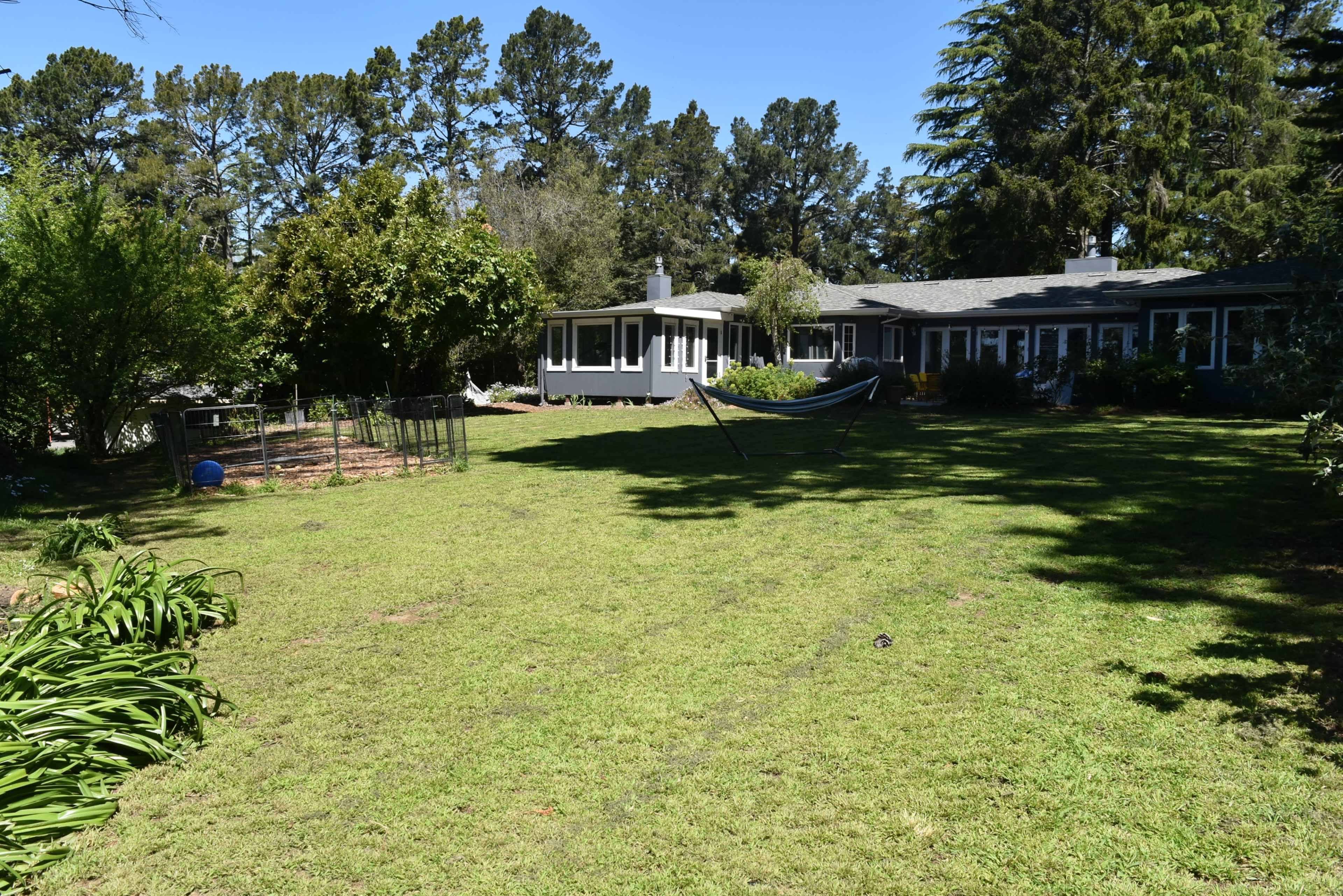 A well-maintained lawn features a hammock between trees, adjacent to a house with large windows and a garden area.