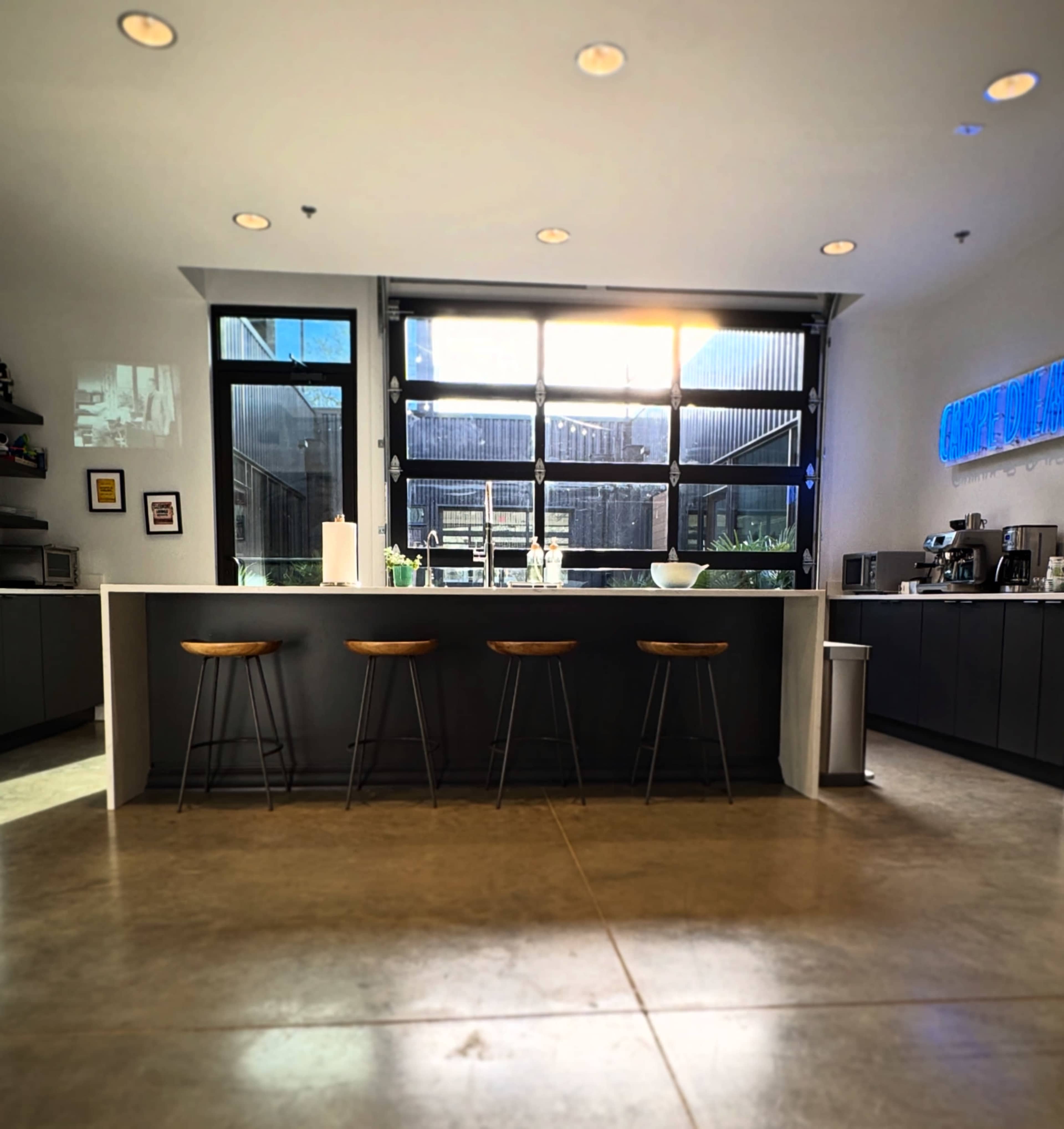 A modern kitchen features a long white countertop with four wooden stools and large windows providing natural light.