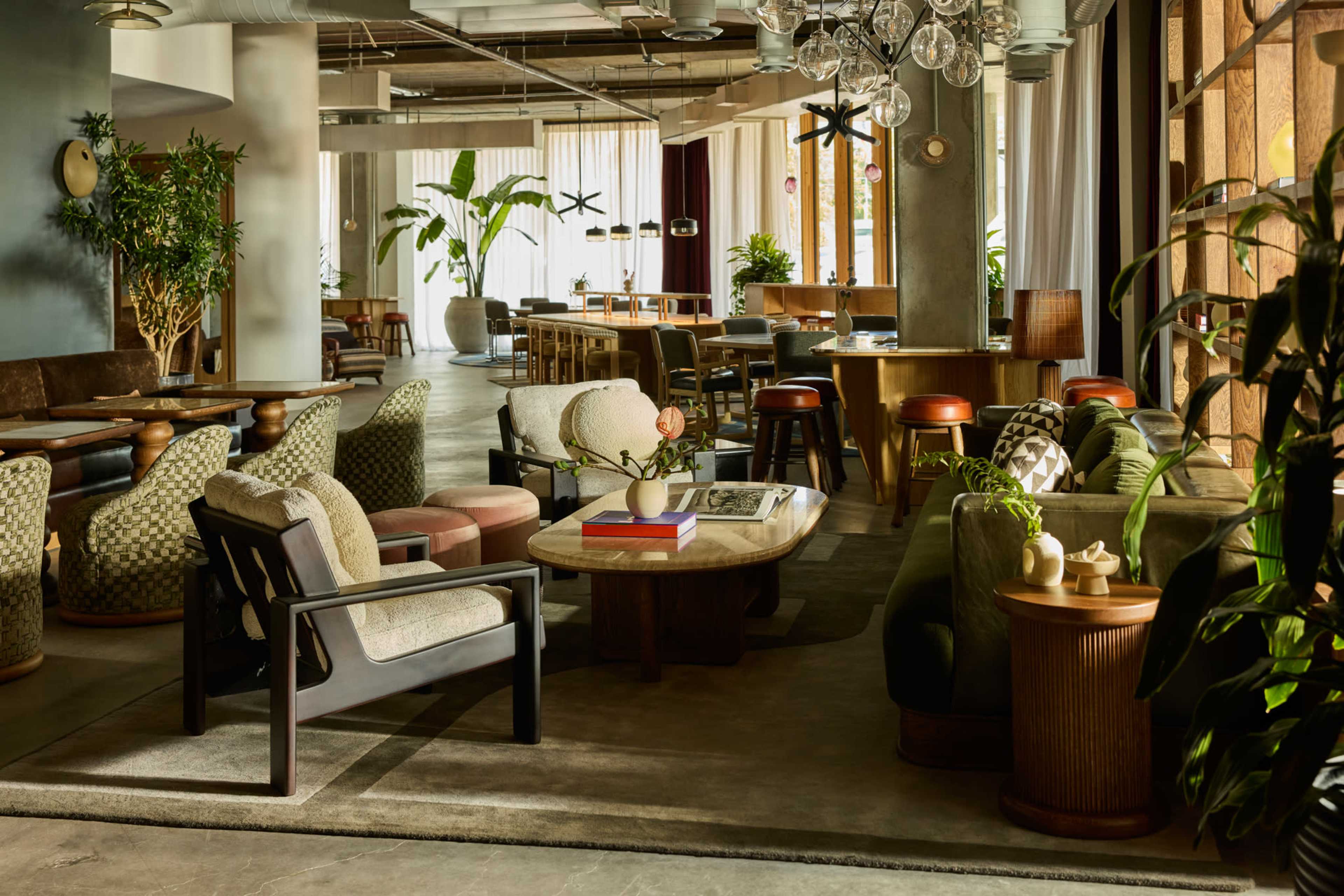 The image depicts a modern, airy café with a mix of seating options, including couches and chairs, surrounded by greenery and large windows letting in natural light.