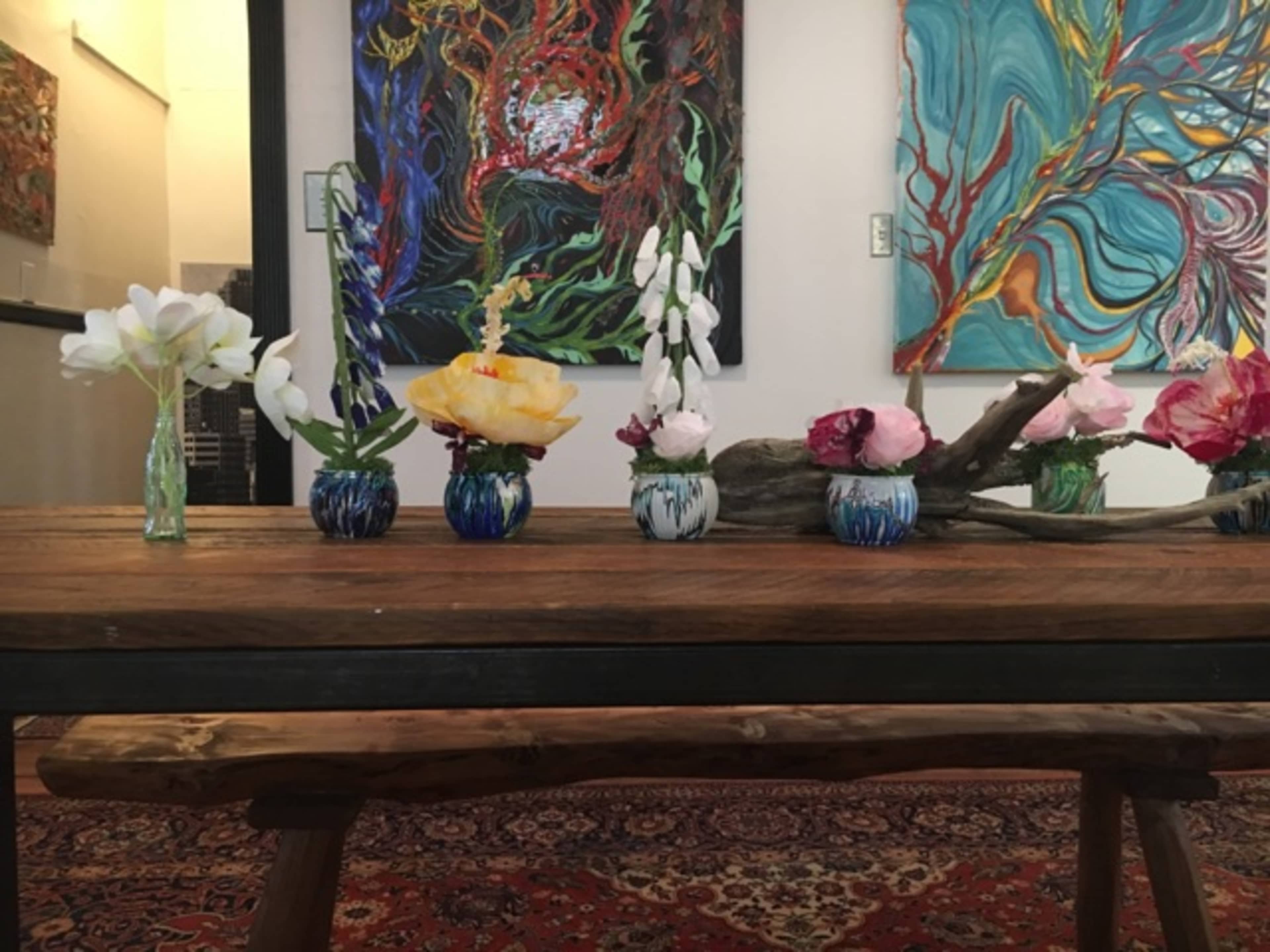 A wooden table adorned with several small vases containing colorful flowers, set against a backdrop of abstract paintings.