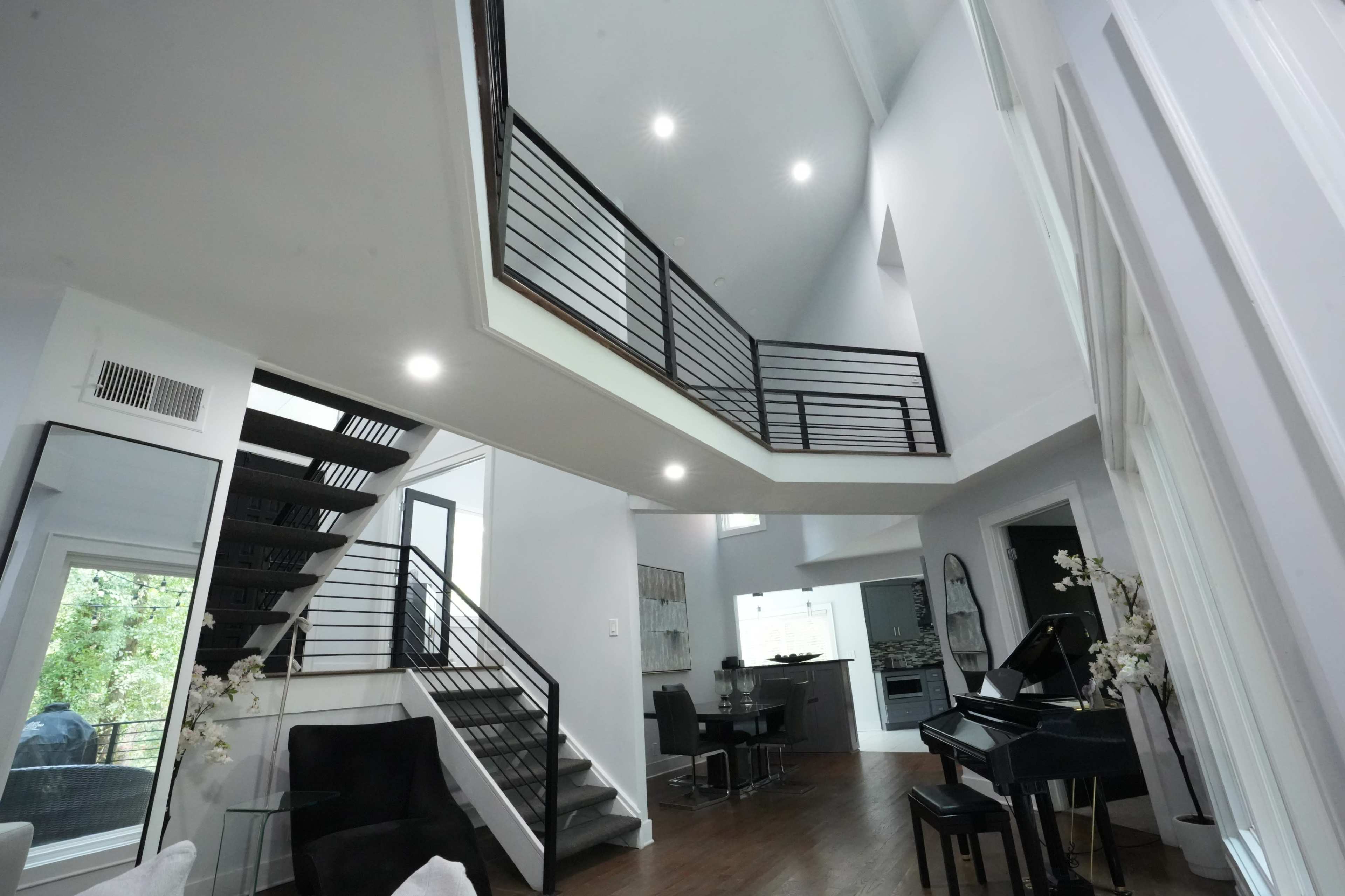 The image shows a modern interior featuring a staircase with black railings, a piano, and an open layout connecting multiple rooms.