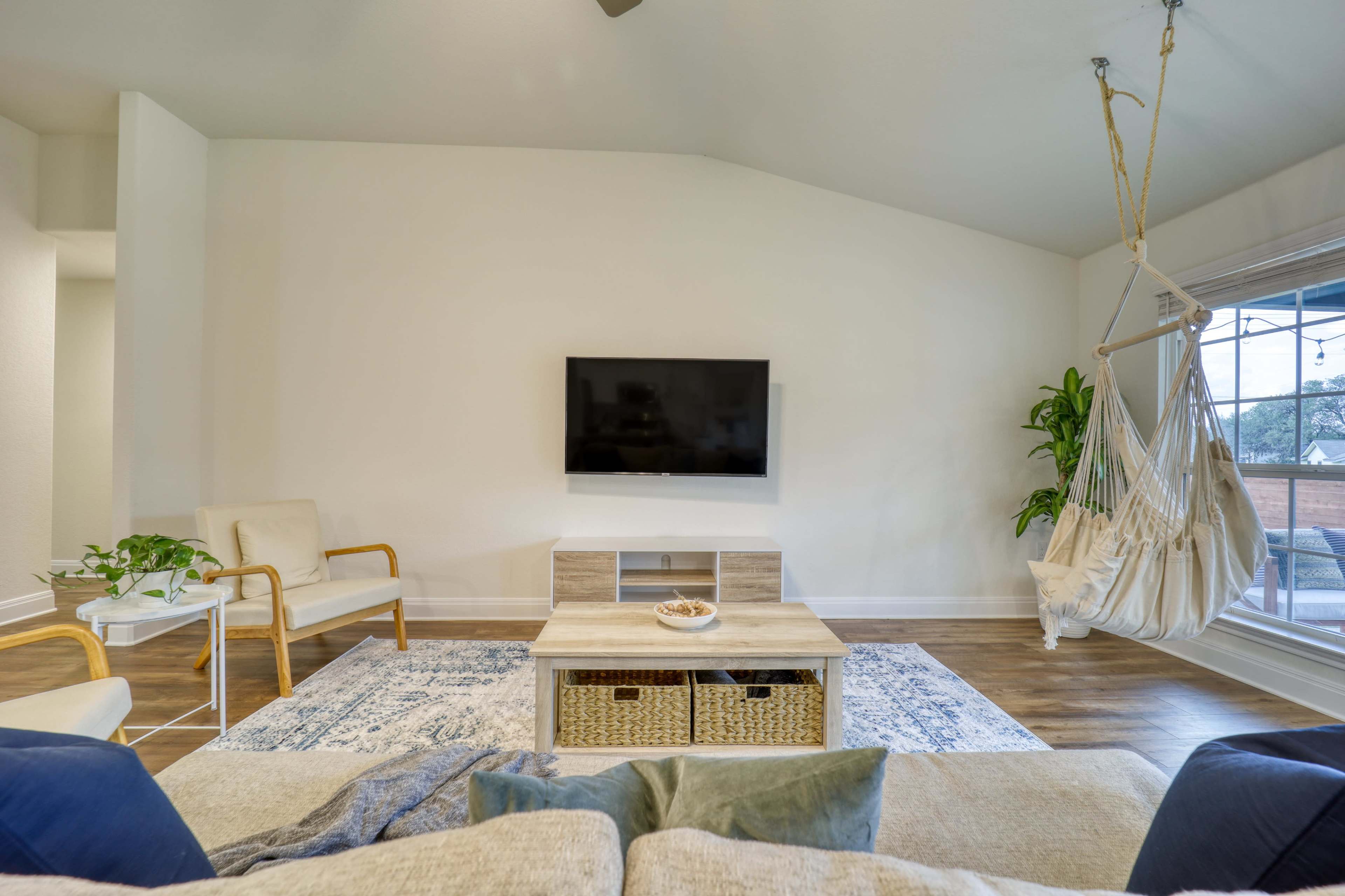 A bright living room with a TV mounted on the wall, a wooden coffee table with decorative items, and a hanging chair beside a window.
