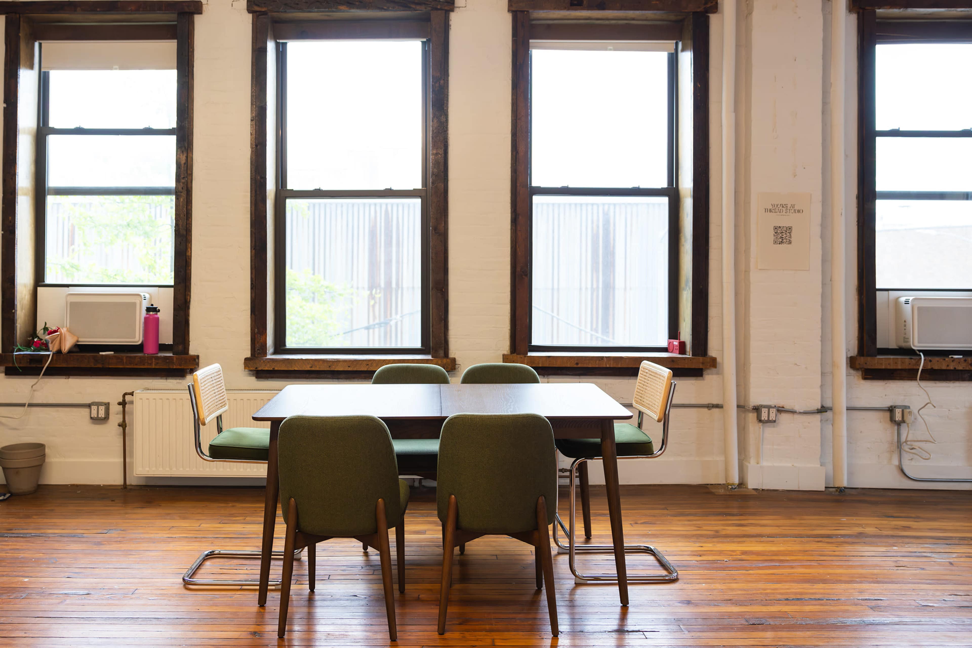 A wooden dining table with green chairs is positioned between three windows in a room with hardwood flooring.