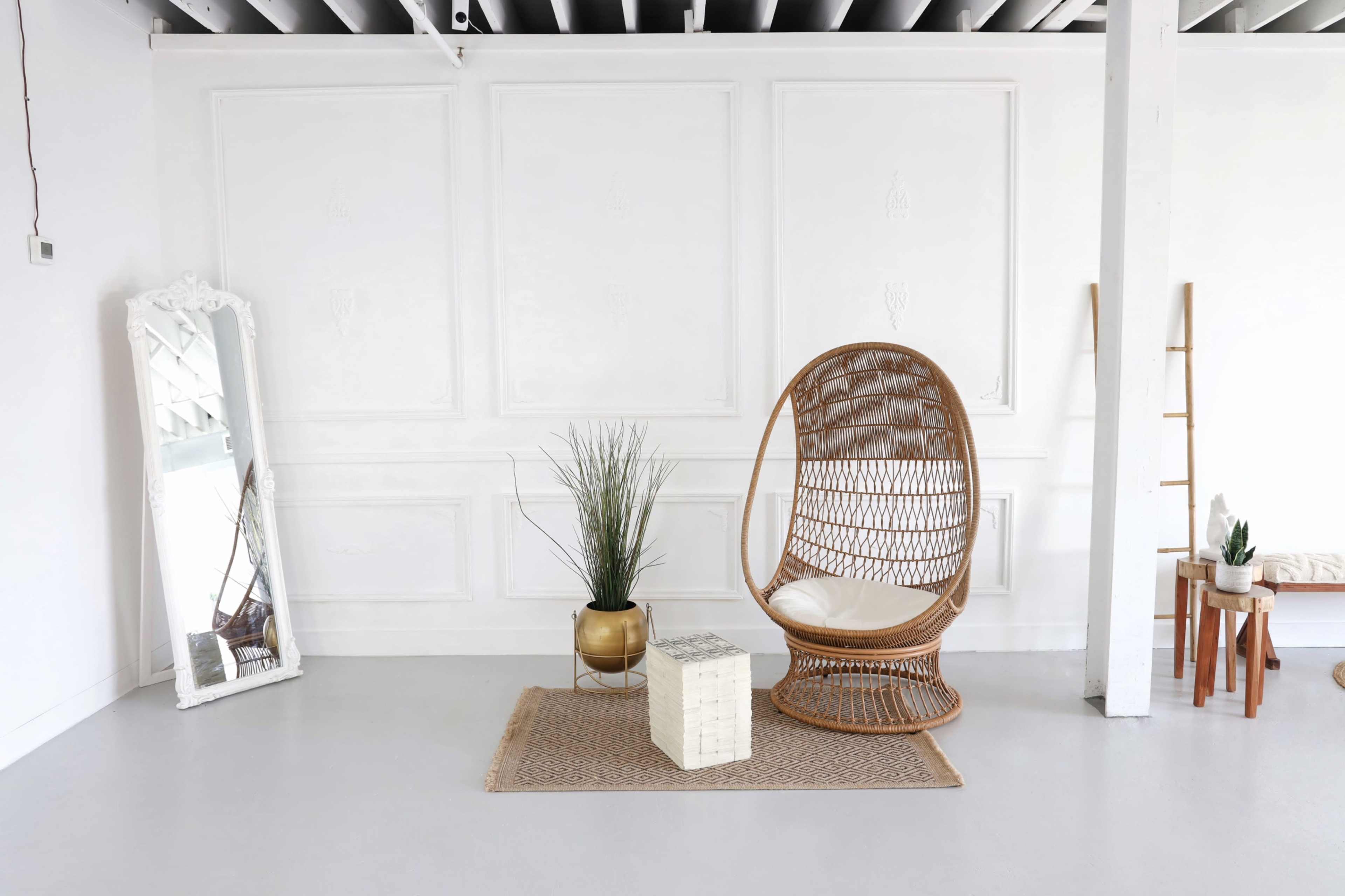 A minimalistic interior space featuring a woven rattan chair, a round gold planter with greenery, a decorative woven rug, a tall mirror, and a small side table.