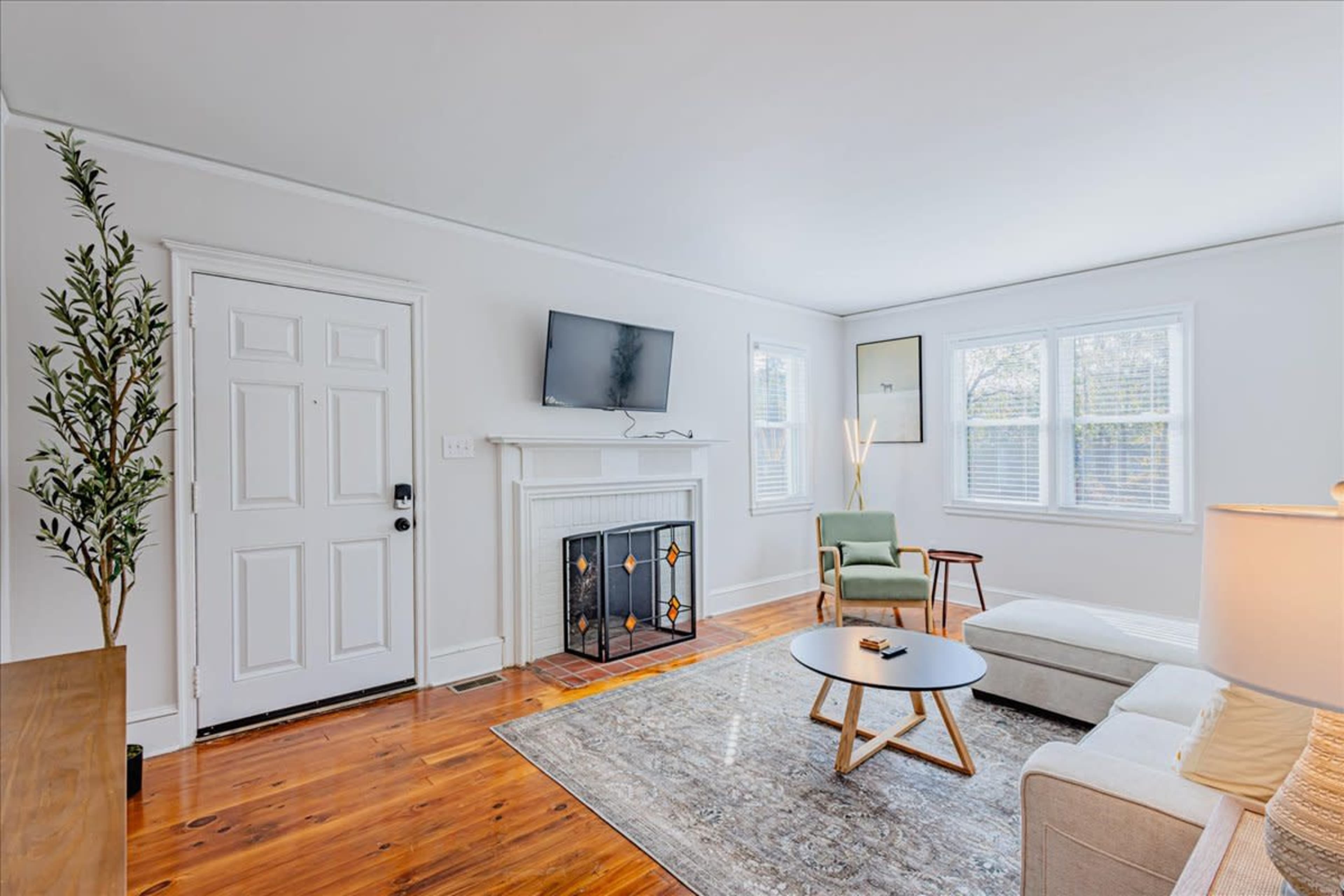 A cozy living room features a fireplace, a mounted television, a coffee table, and a sectional sofa, with large windows allowing natural light.