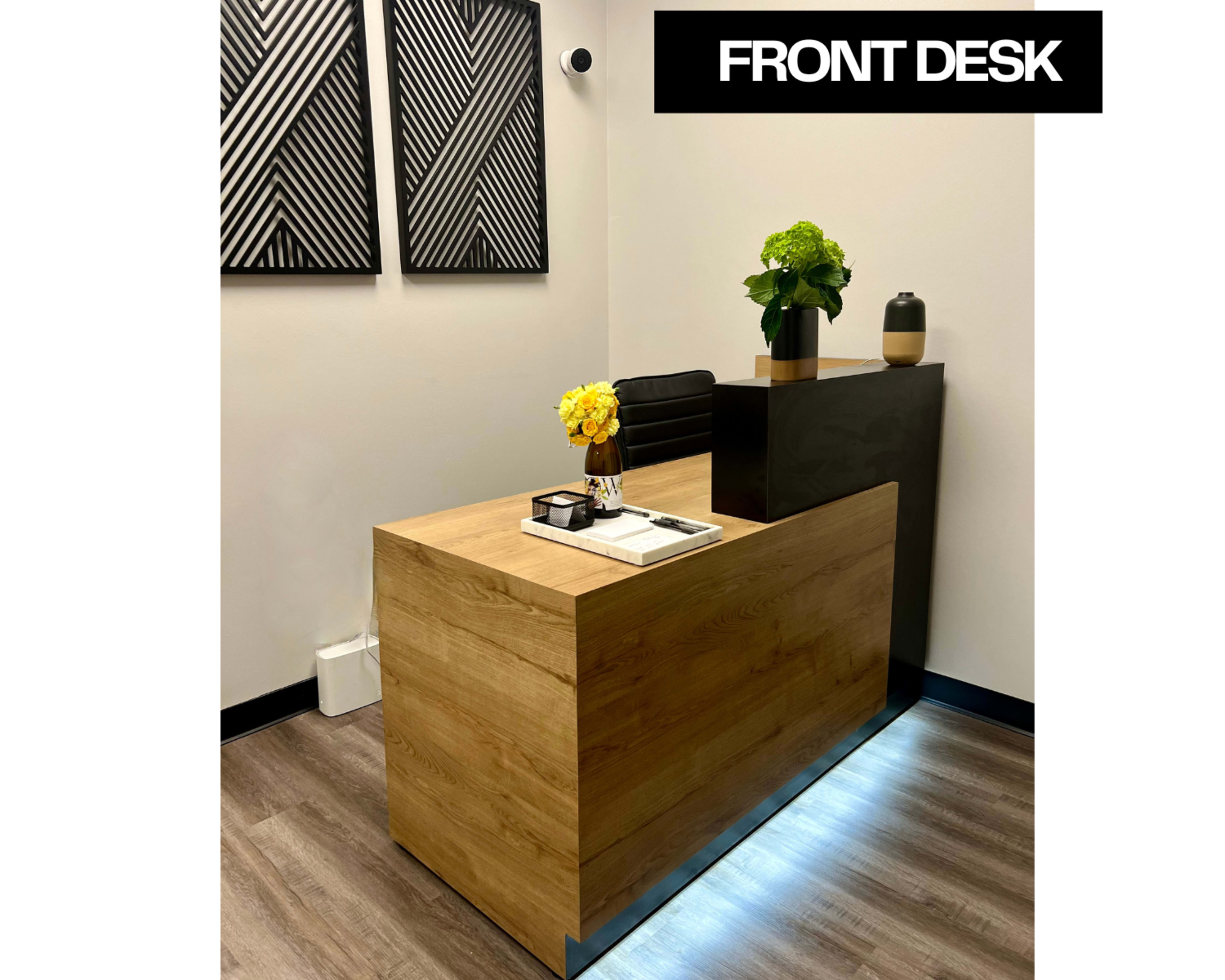 The image shows a modern front desk made of wood, featuring a small vase of flowers and decorative items on top, against a backdrop of geometric wall art.