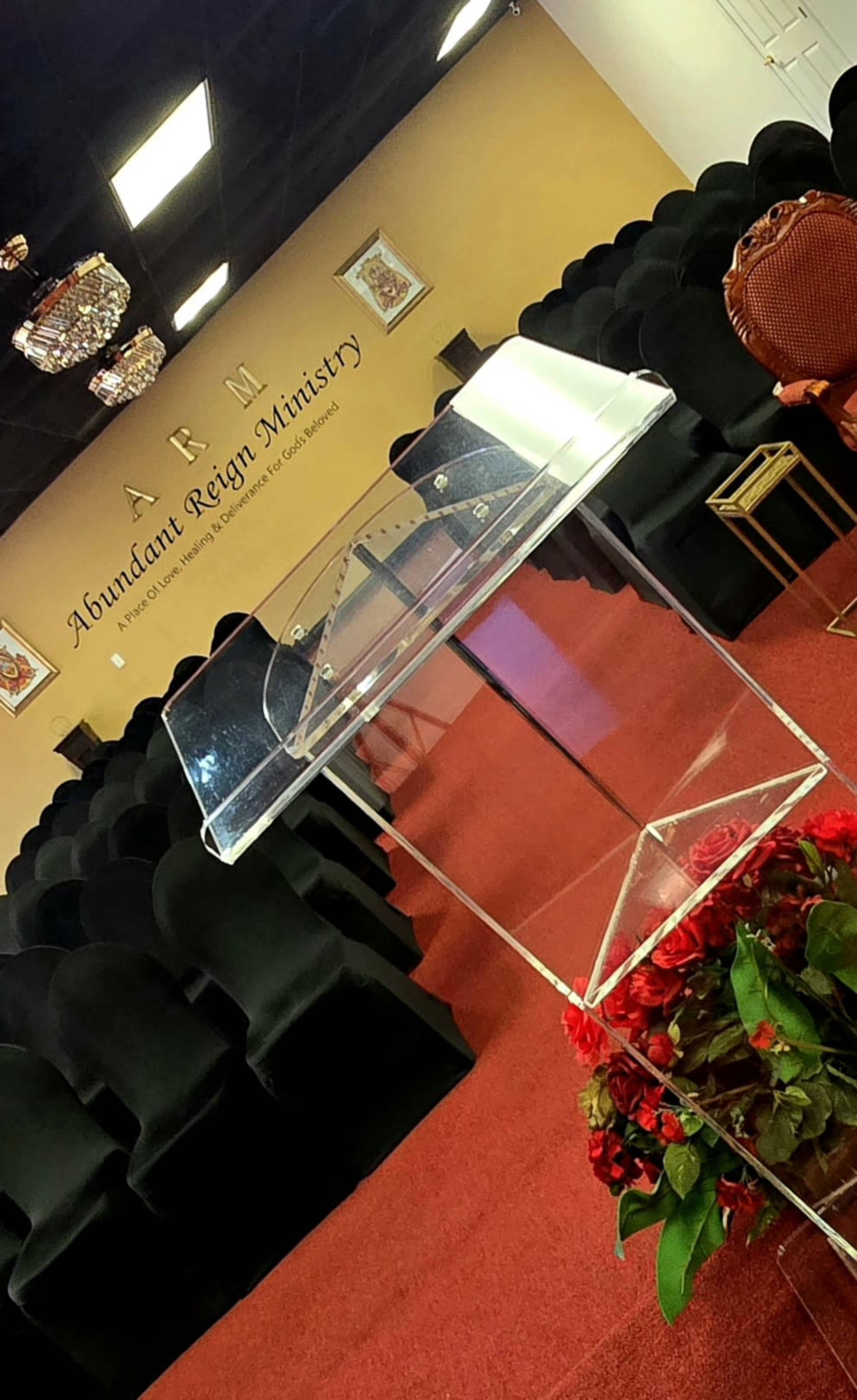 The image shows a pulpit adorned with flowers in a church sanctuary, surrounded by rows of black chairs.
