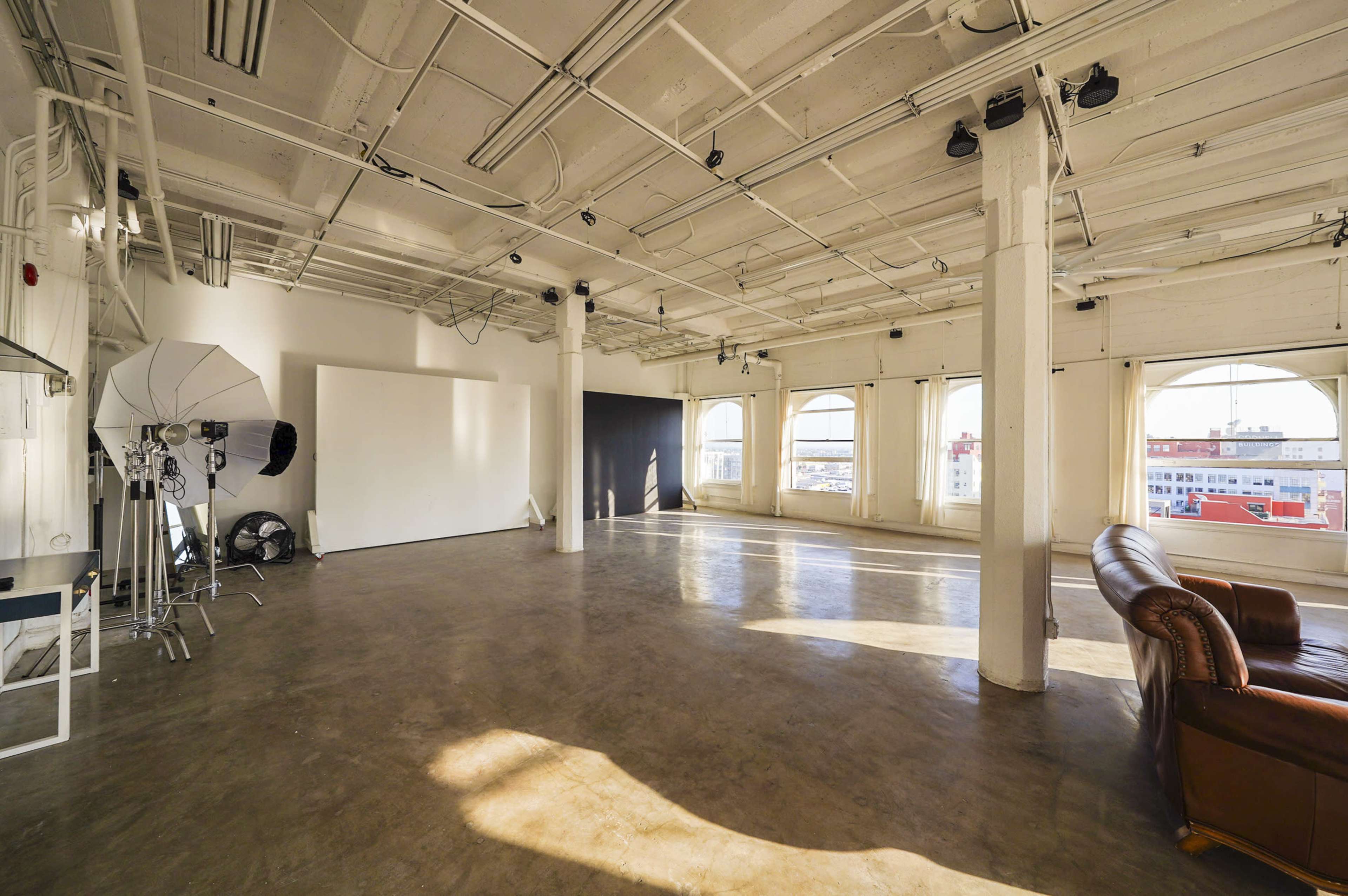 A spacious studio with large windows, a concrete floor, and a brown leather couch, along with studio equipment on one side.