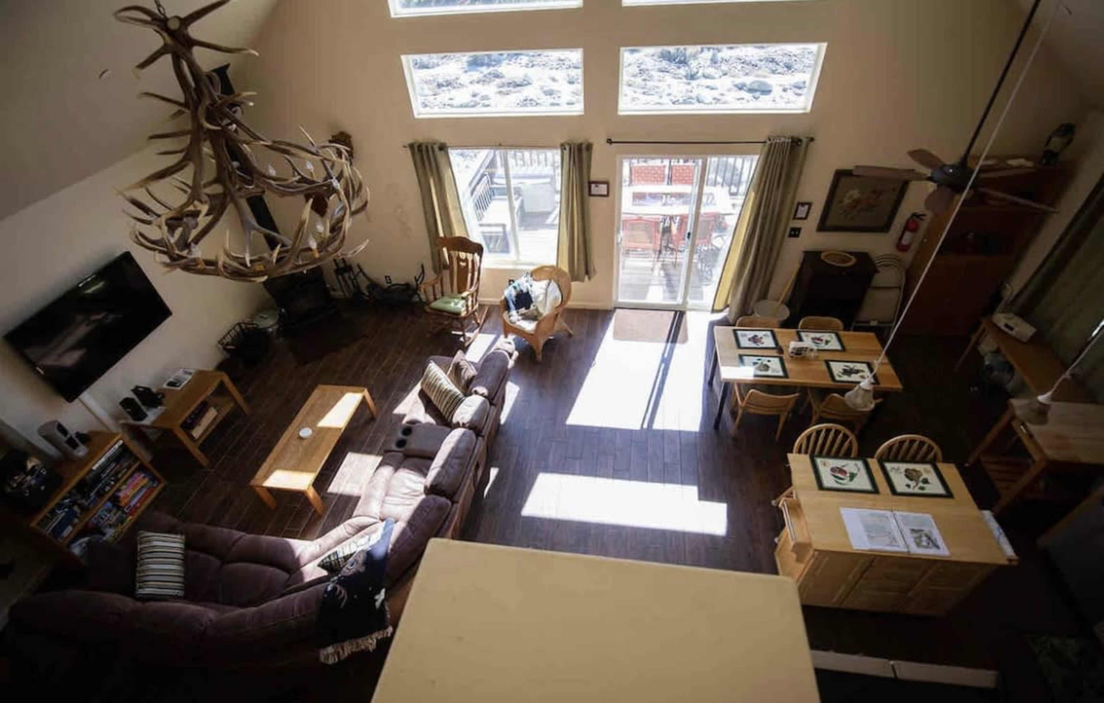 A spacious living area with high ceilings, featuring a large window, a dining area with wooden tables and chairs, and a seating arrangement centered around a television.