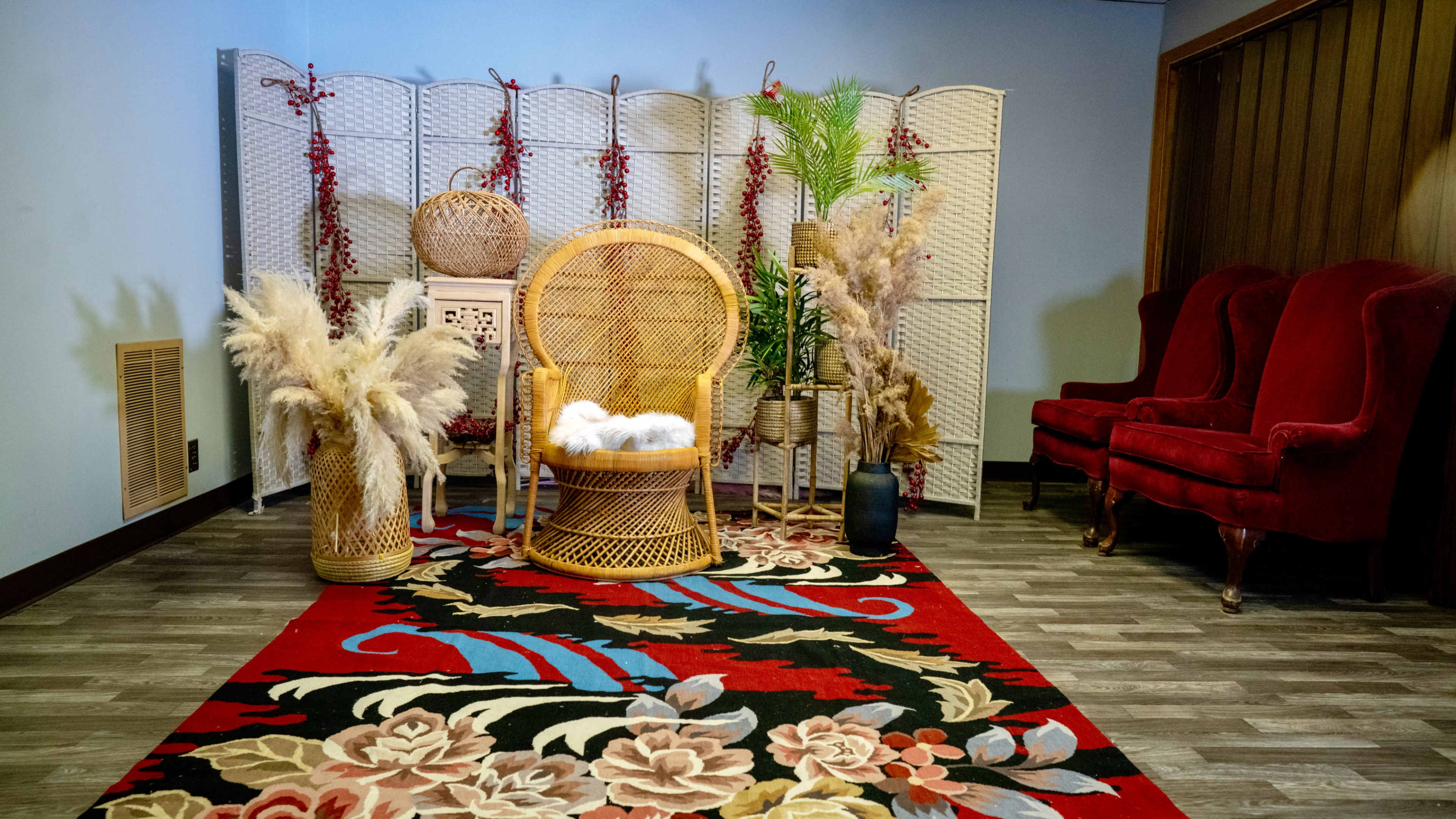 A cozy interior setup featuring a rattan chair, decorative plants, and red accent chairs, arranged on a floral patterned rug.