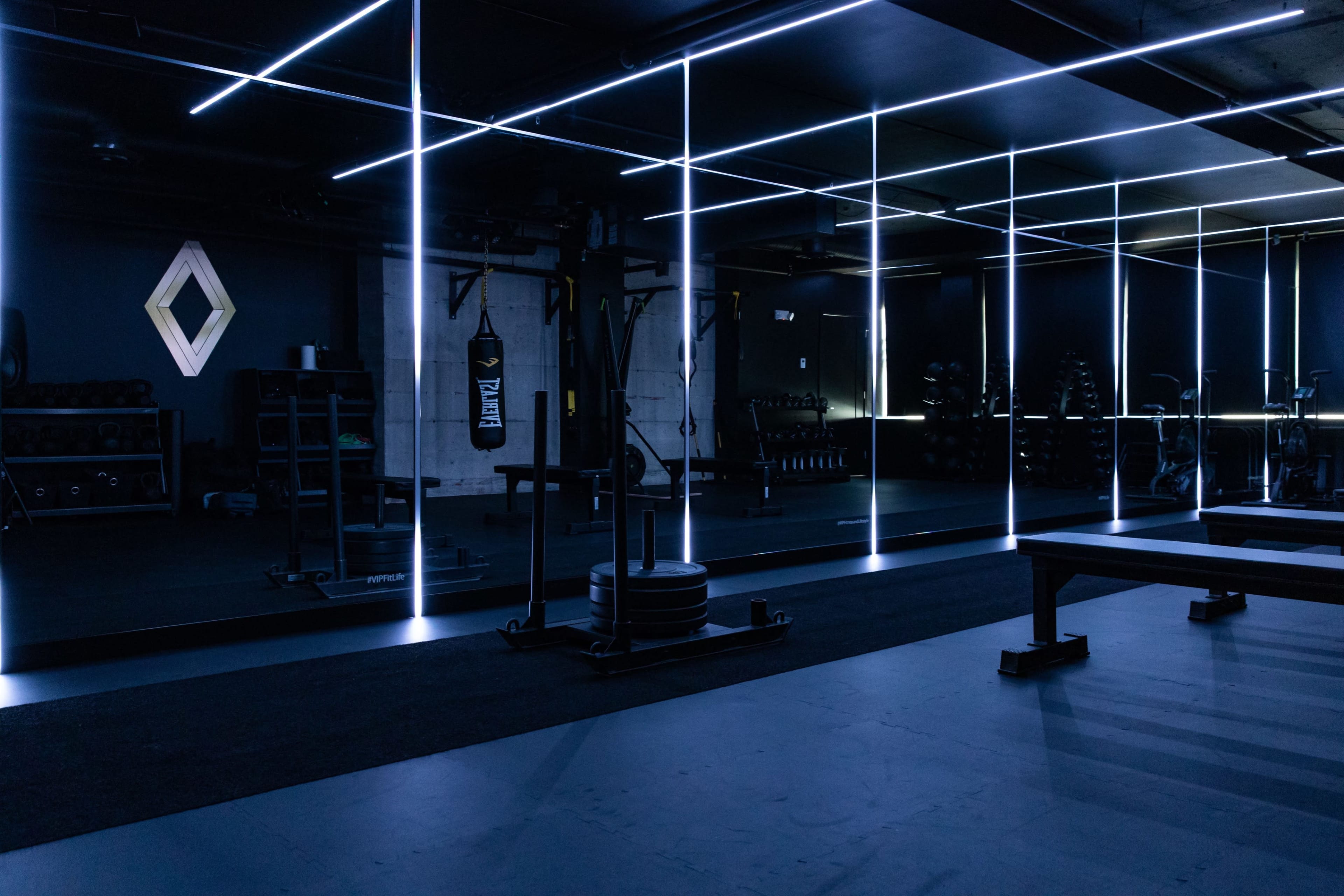 A modern gym features dark walls, illuminated lines, weightlifting equipment, and a mirrored area.