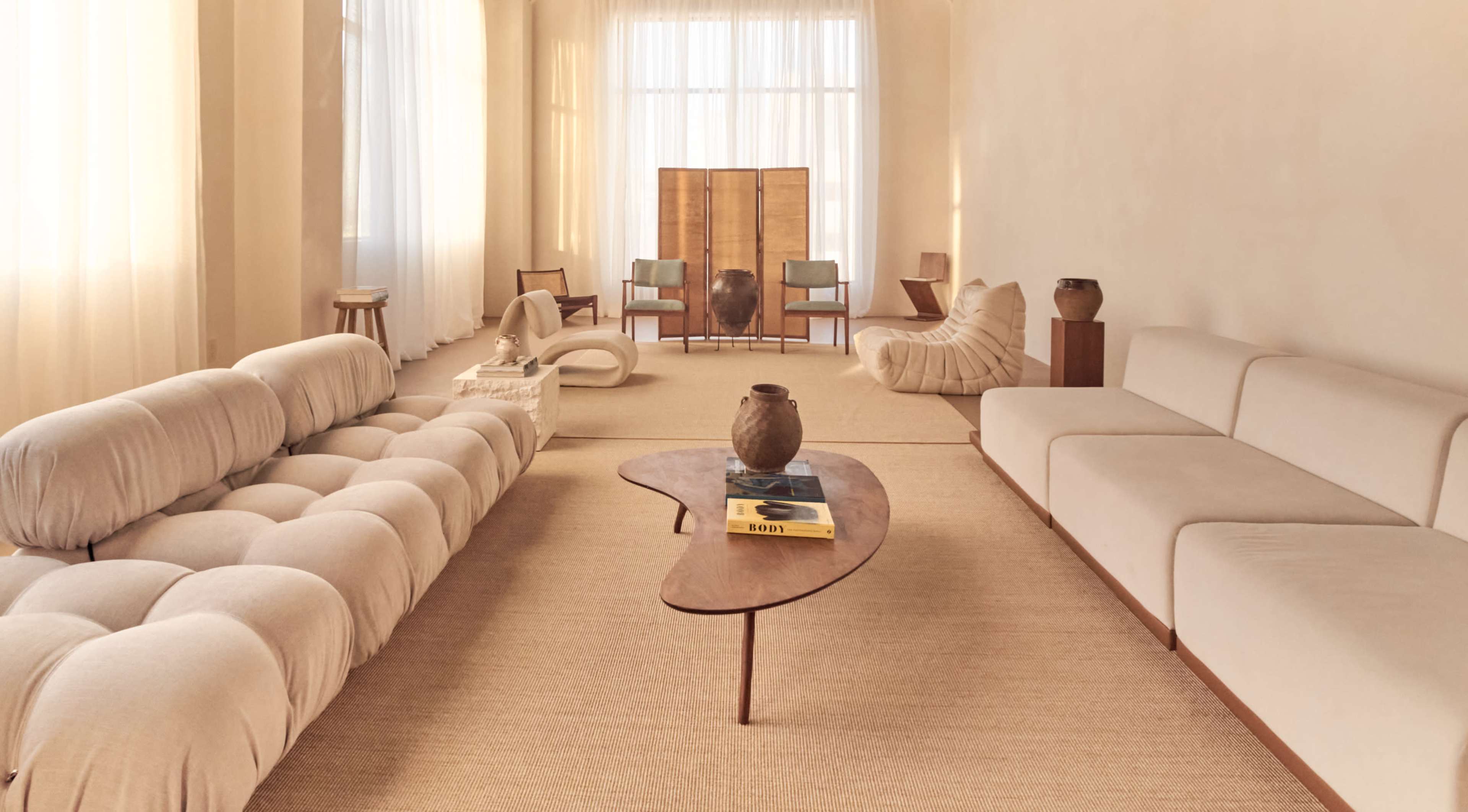 The image depicts a minimalist living room featuring a curved sofa, two modern chairs, and a wooden coffee table, all arranged in a space with large windows and natural light.