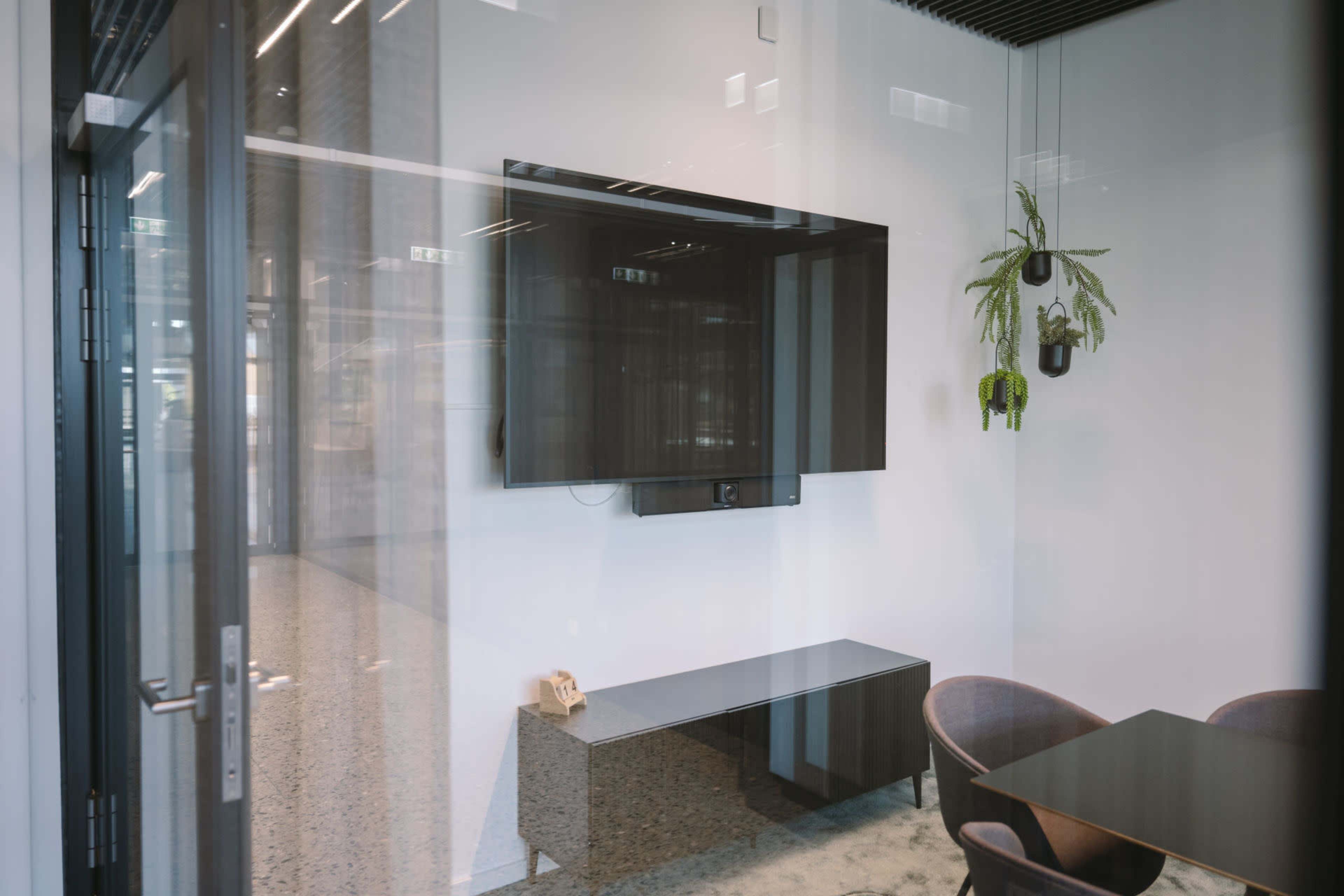 A modern conference room features a wall-mounted television, a sleek table, and chairs, with a plant hanging in the corner.