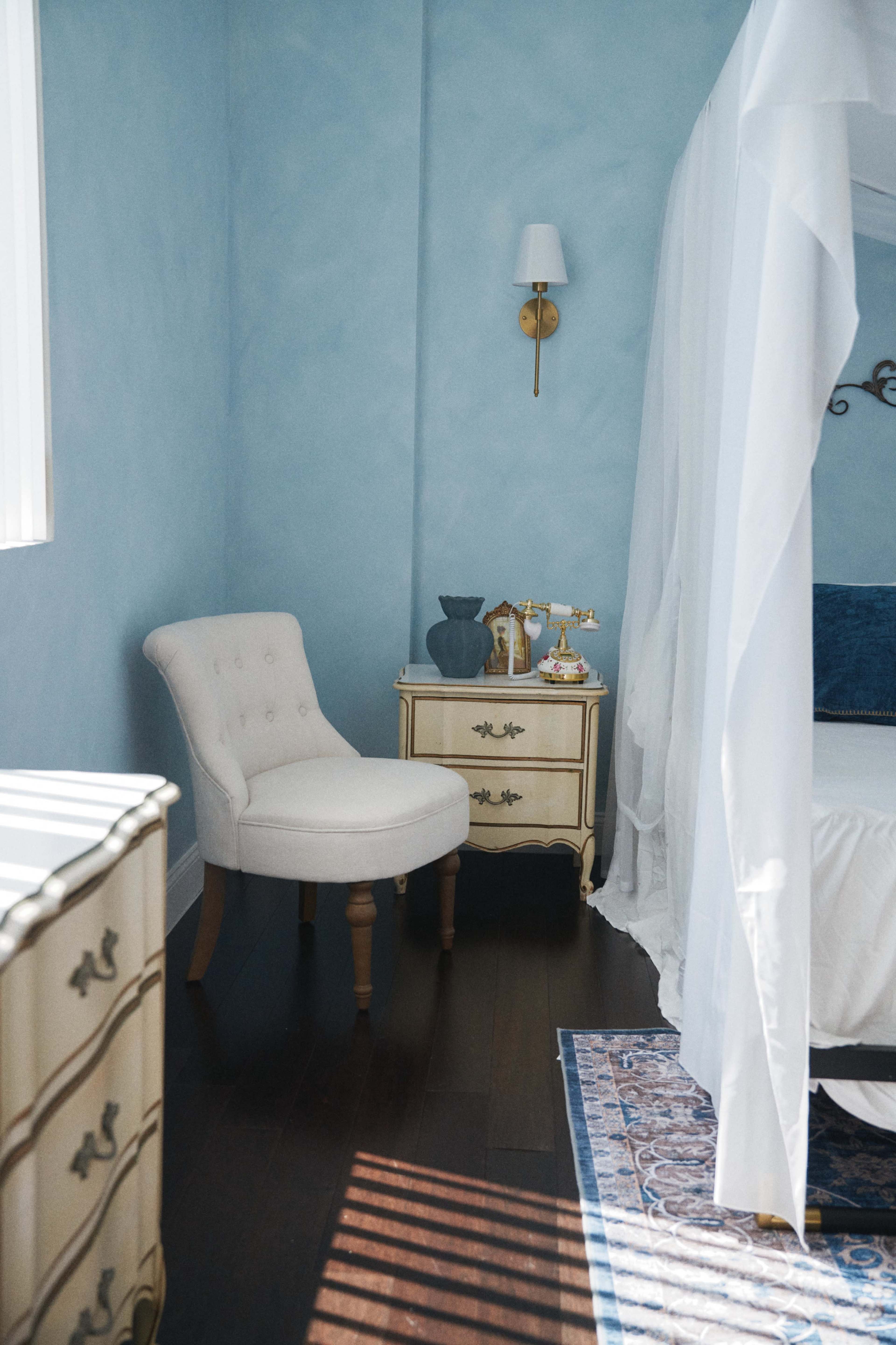The image shows a softly lit bedroom with a blue wall, a white canopy bed, a small nightstand adorned with decorative items, and a chair nearby.