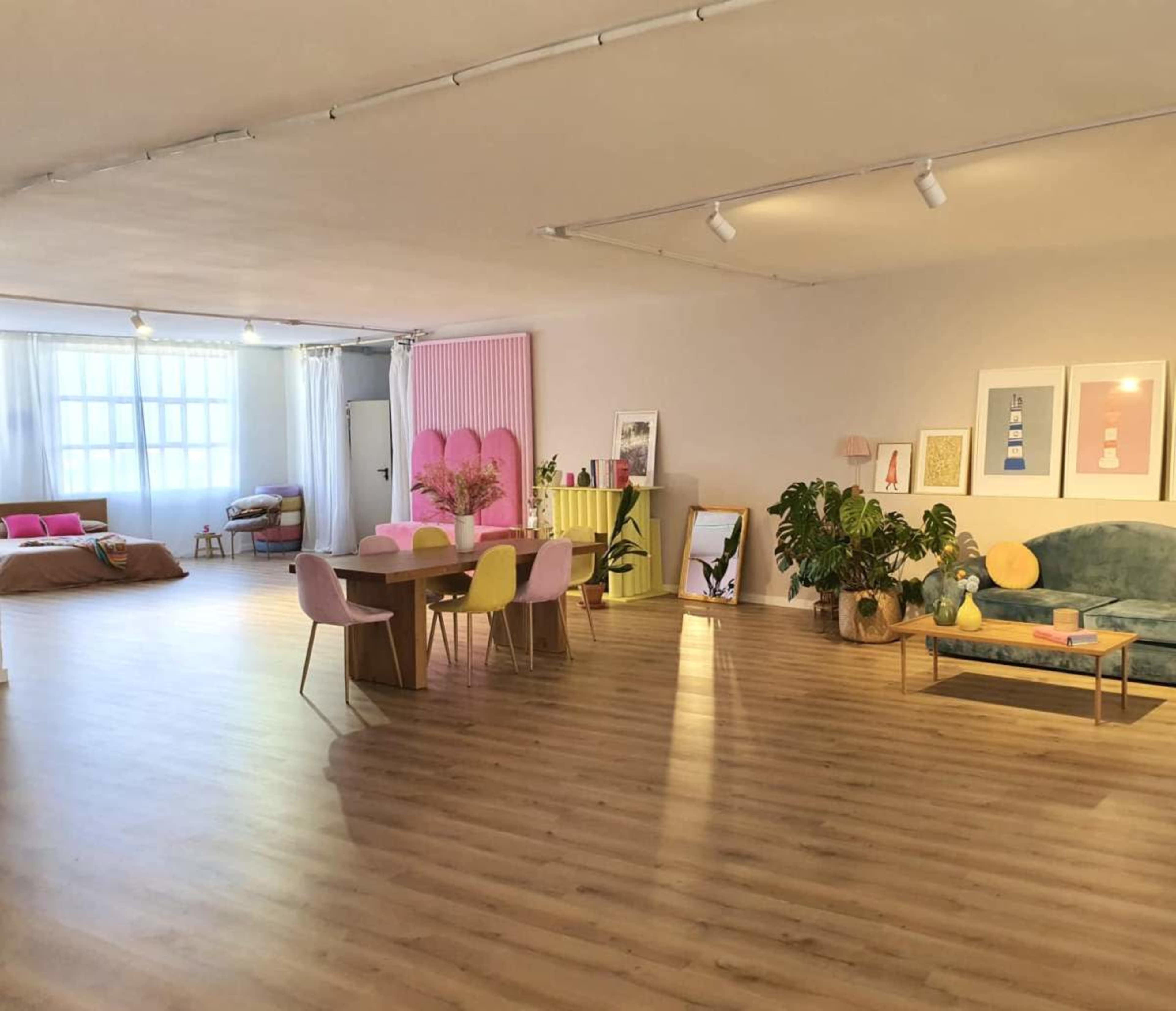 The image shows a spacious, brightly lit room with wooden flooring, featuring a dining table with multicolored chairs, a green couch, plants, and various decorative items on display.
