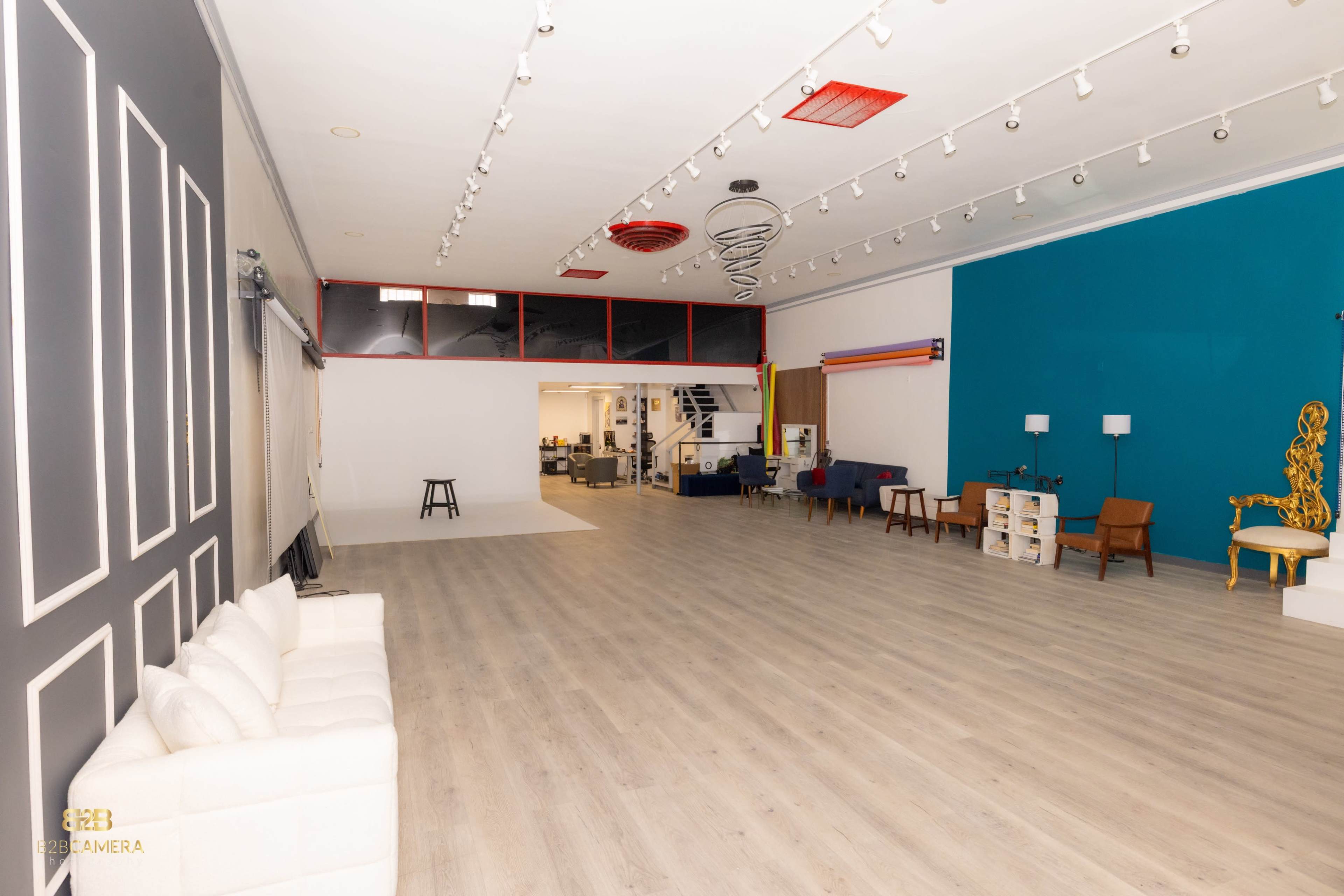 The image shows a spacious, empty room with wooden flooring, white walls, and a colorful accent wall on one side, featuring various furniture pieces such as sofas, tables, and chairs.