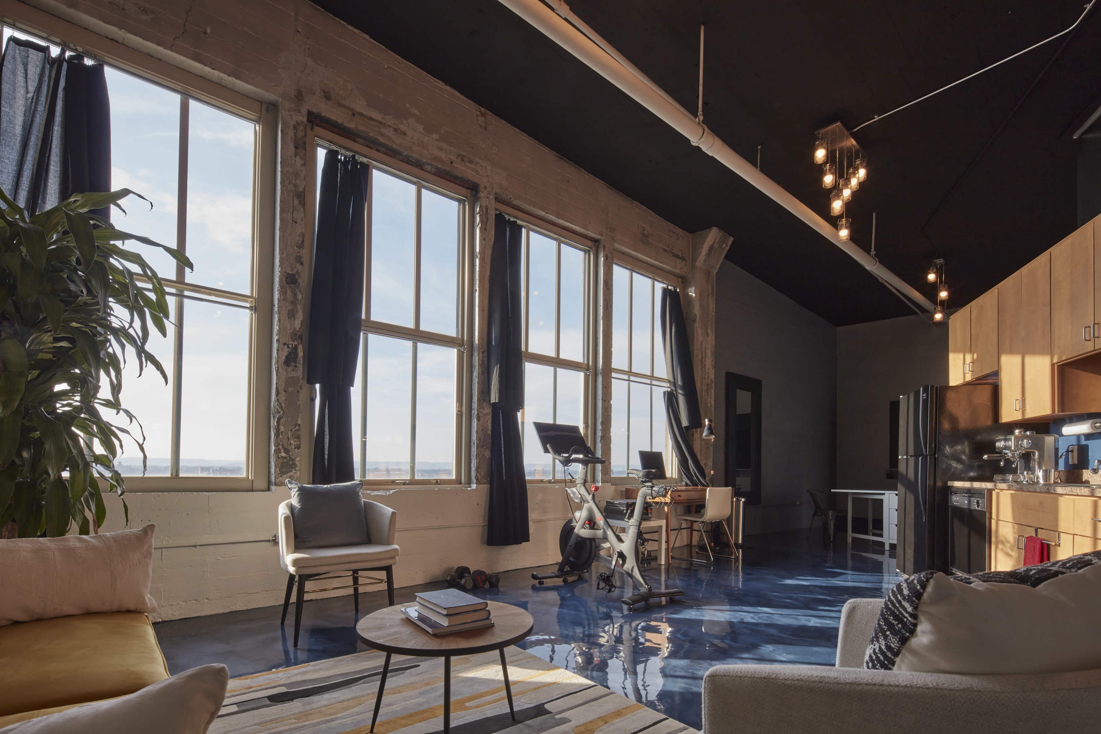 An industrial-style living space with large windows, a workout bike, a kitchen area, and modern furniture.