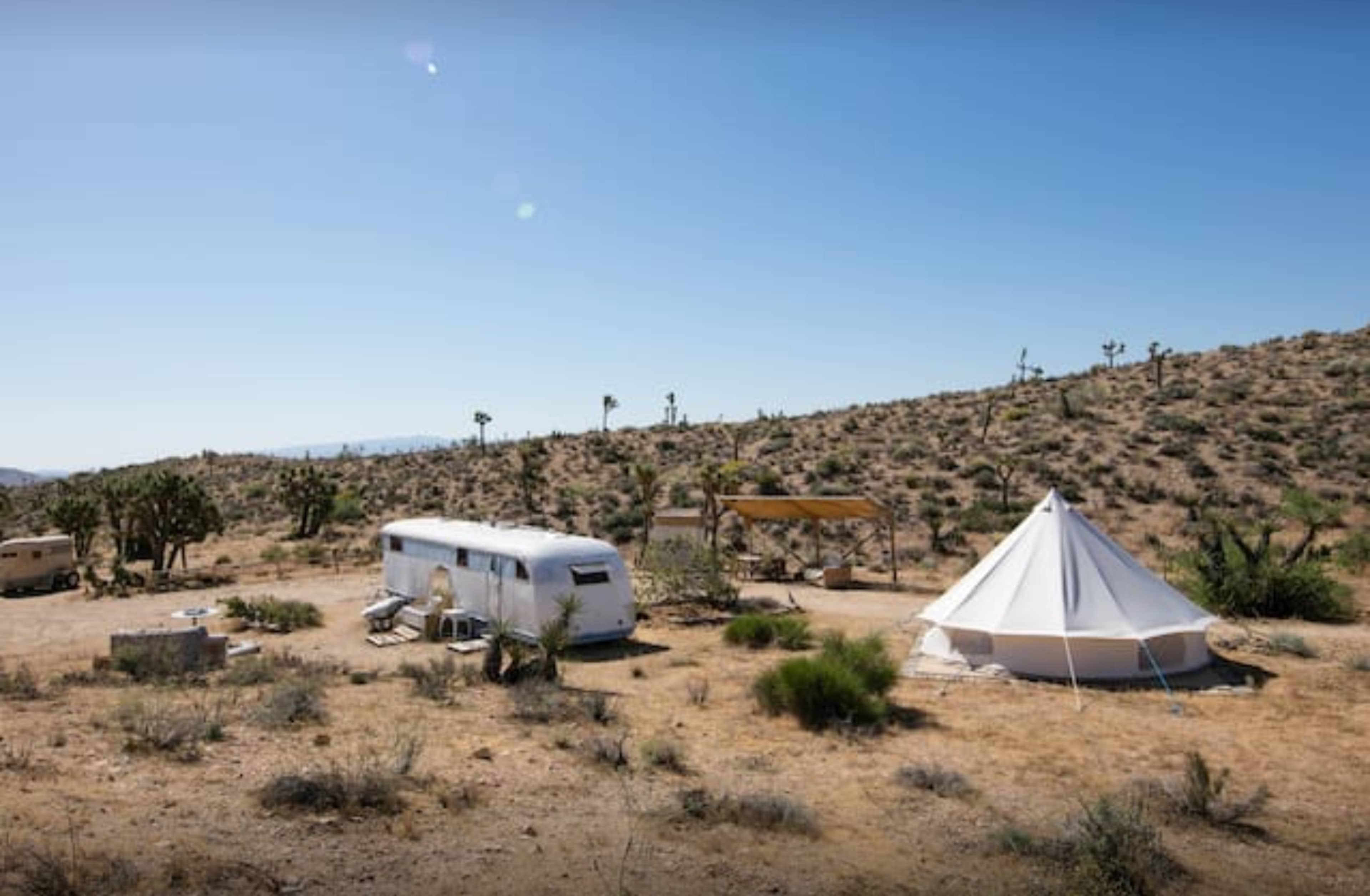 A white vintage trailer is parked next to a large white tent in a desert landscape with sparse vegetation and distant hills.