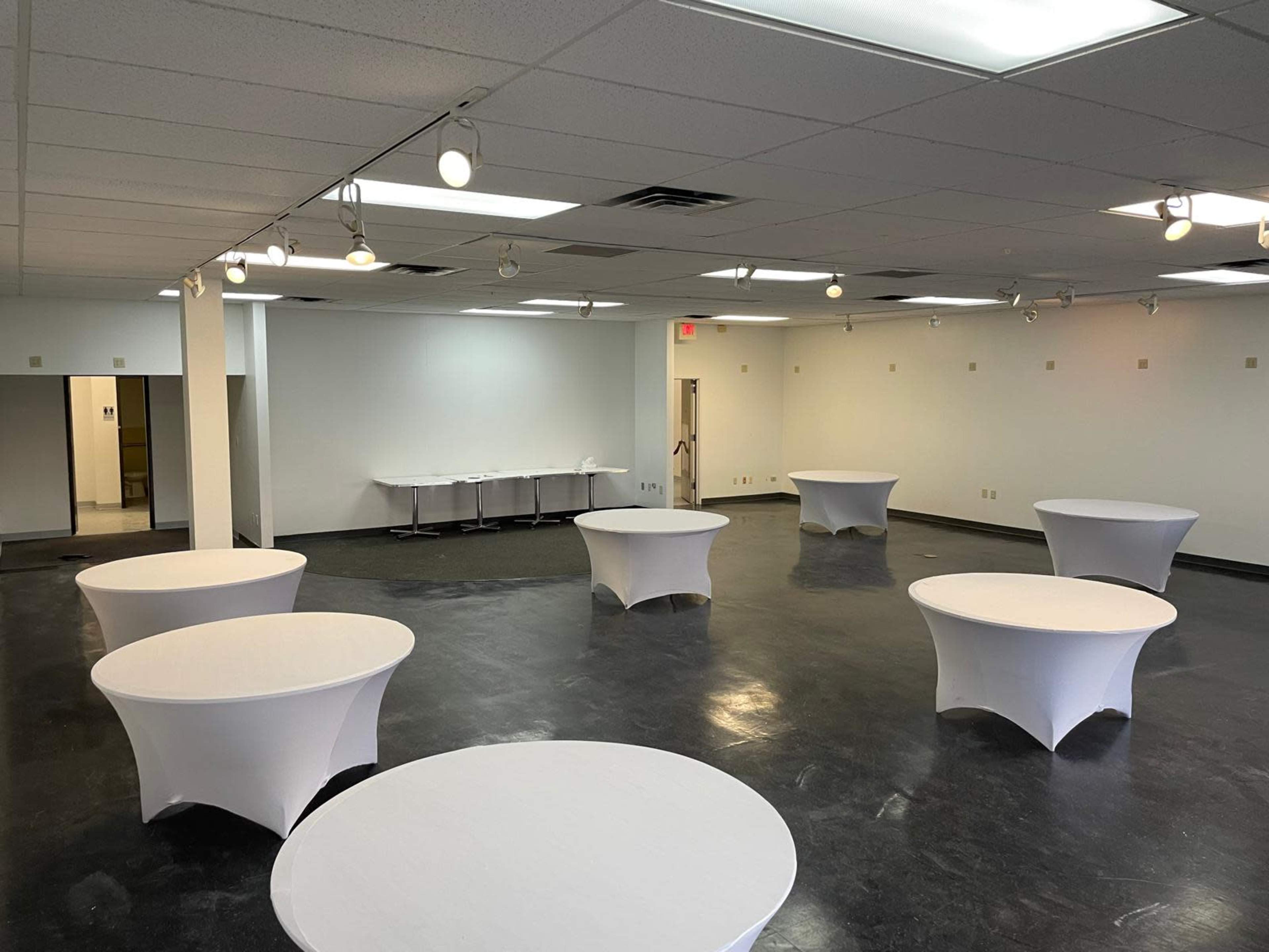 The image shows a spacious, empty room with several round tables covered in white tablecloths arranged throughout.