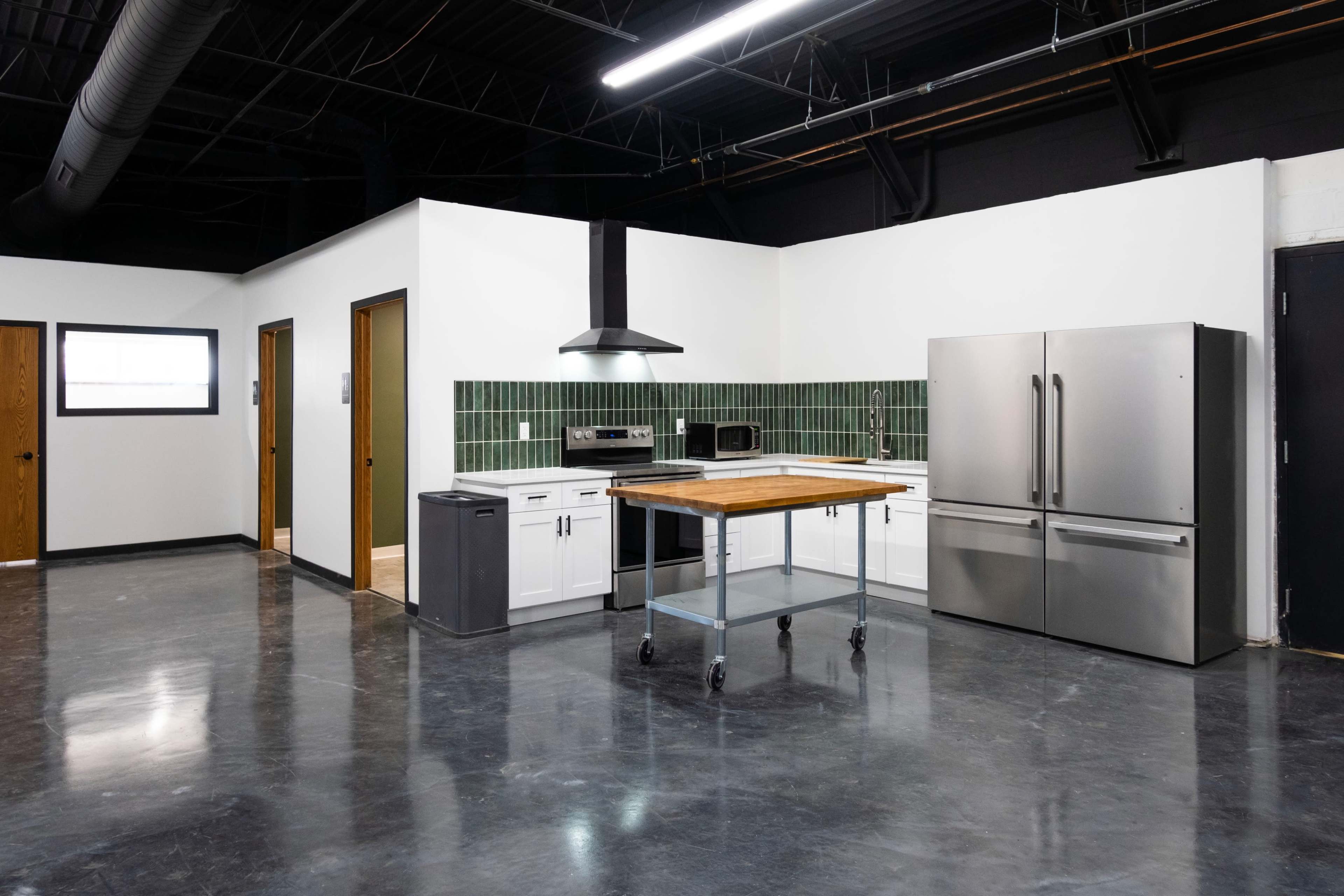 A modern kitchen with stainless steel appliances, a wooden island on wheels, and green tiled backsplash, set in an open space with polished concrete flooring.