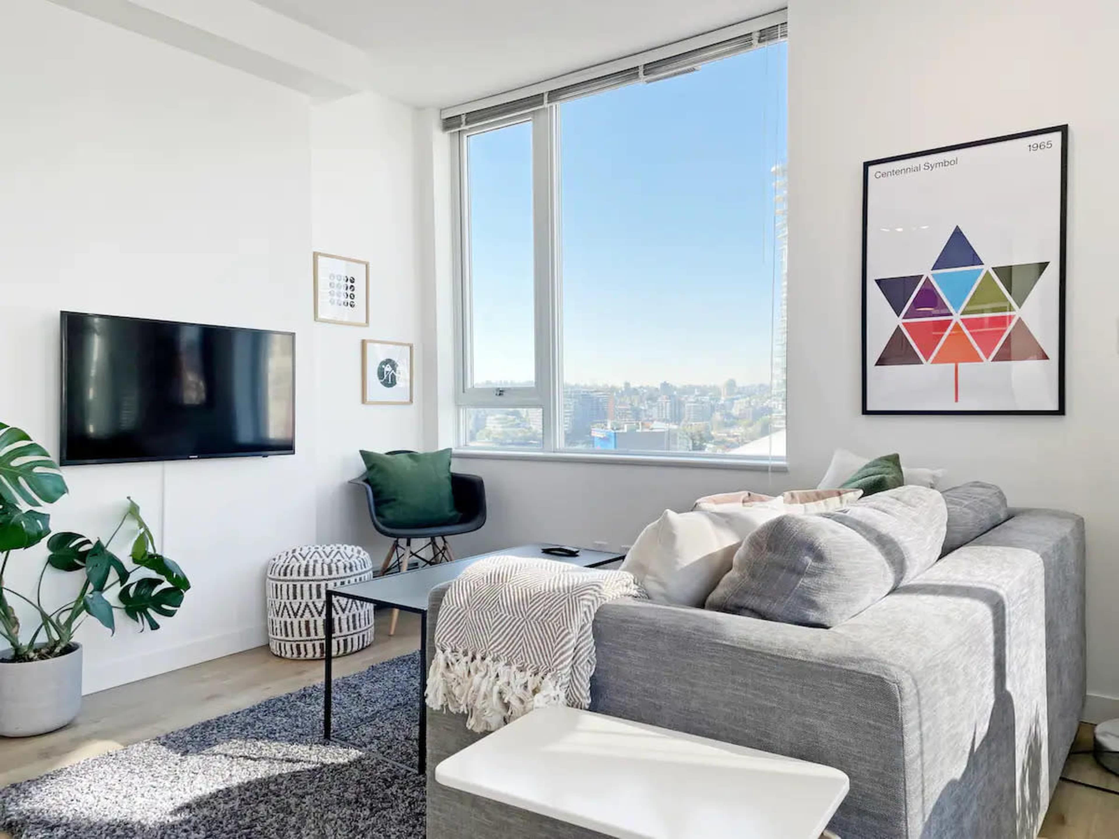 A modern living room features a gray sectional sofa, a black coffee table, a wall-mounted television, and large windows that provide a view of the city skyline.