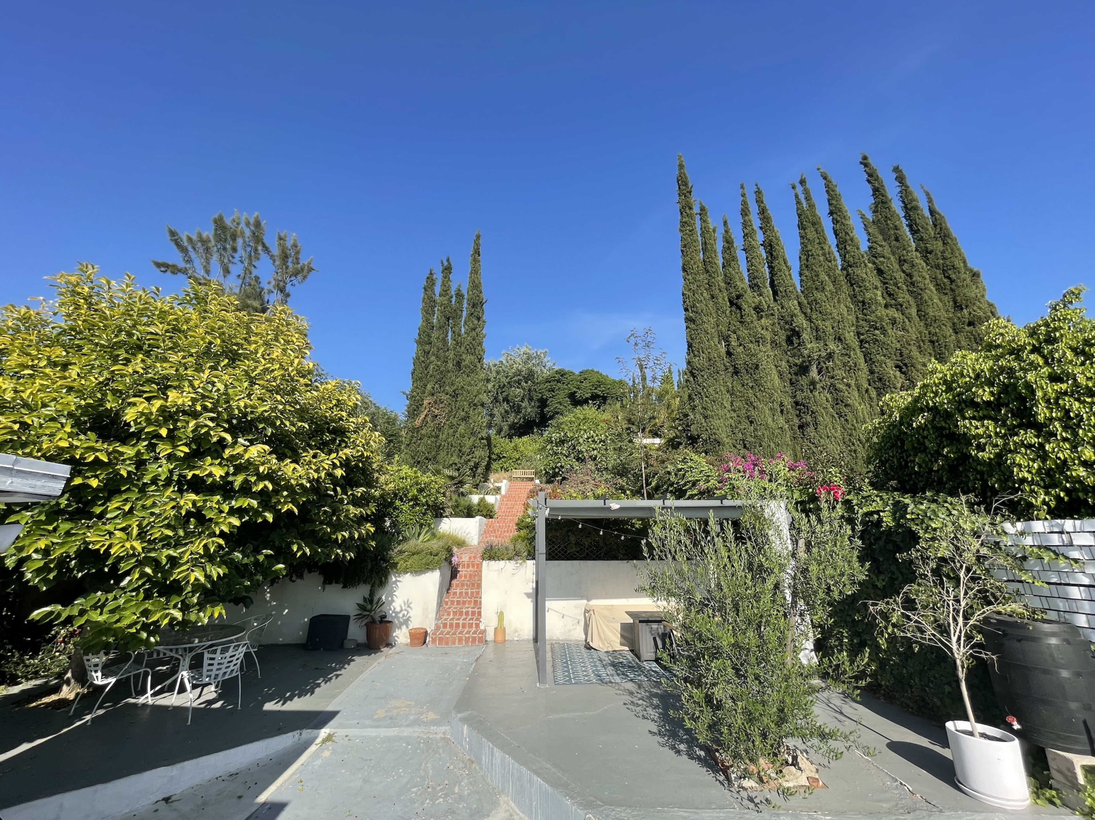 A staircase leads up through a landscaped garden featuring tall cypress trees and various plants under a clear blue sky.