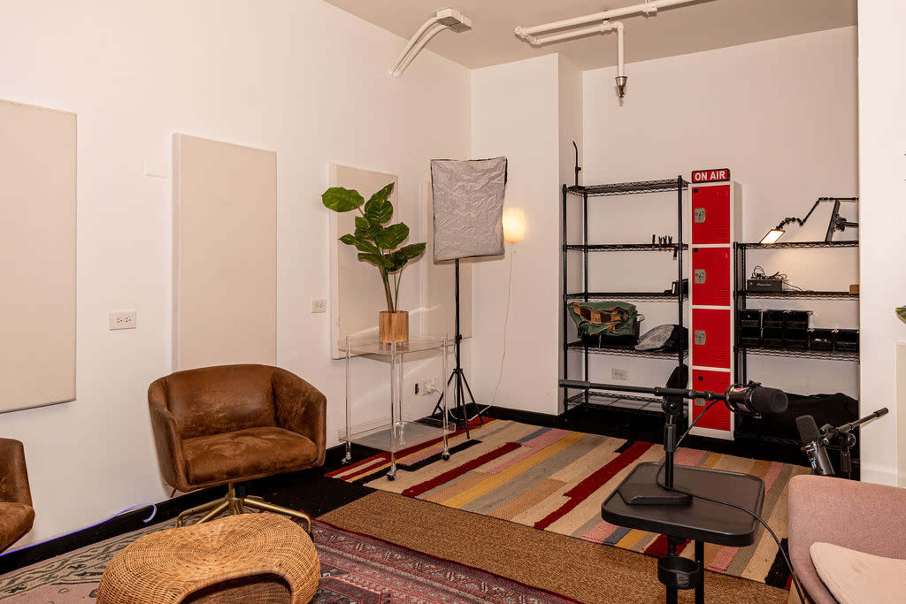 The image depicts a small, modern studio space featuring two brown chairs, a shelving unit with various equipment, a light source, and acoustic panels on the walls.