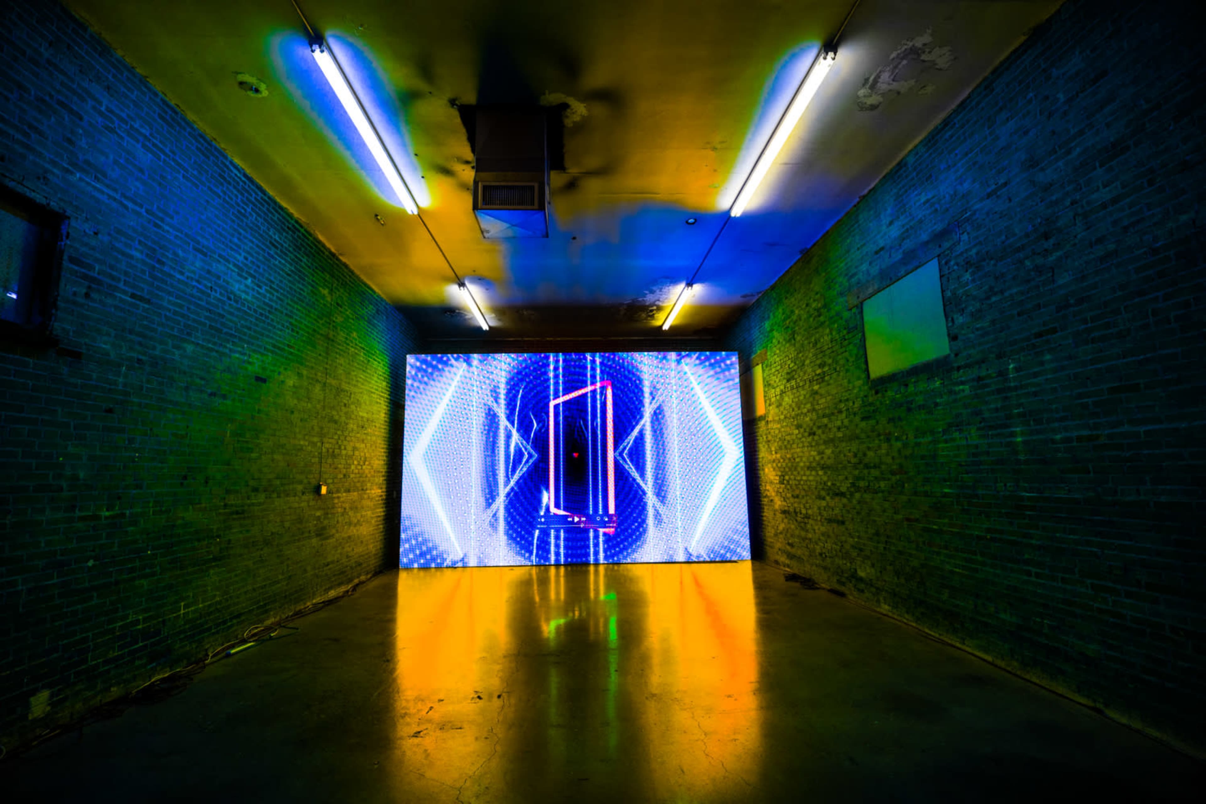 A dimly lit room features a large, brightly illuminated screen displaying dynamic visual patterns on the far wall.