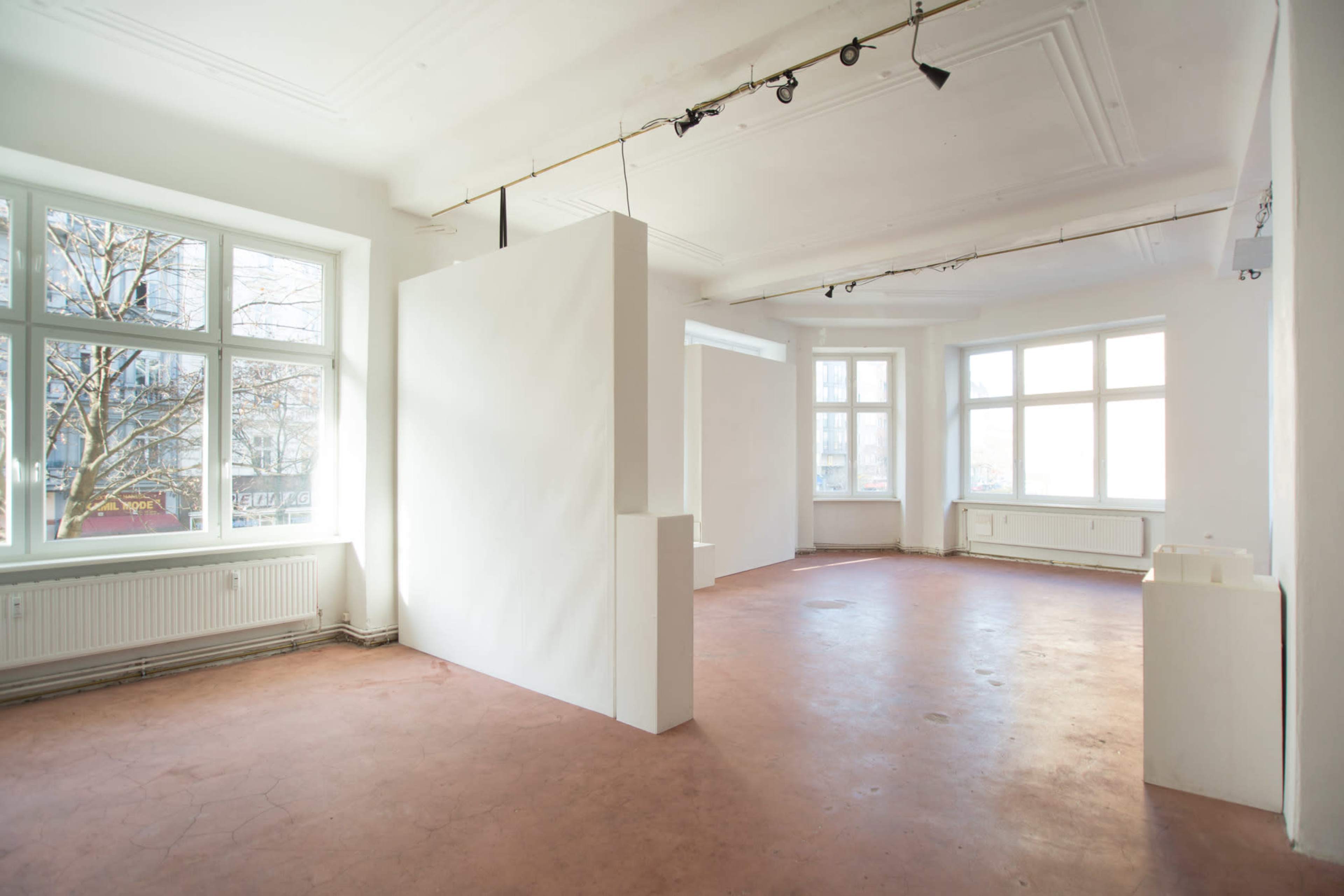 A spacious, unfurnished room with white walls and large windows, featuring a bare concrete floor and a few partition walls.