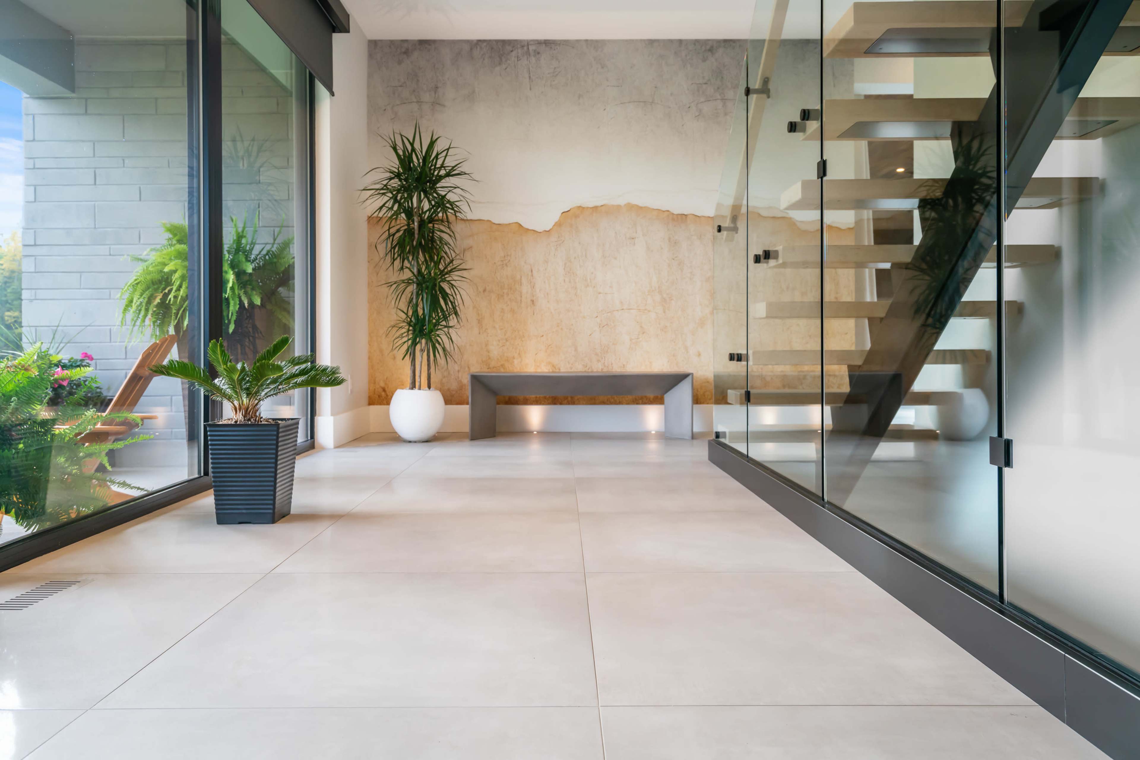 A modern interior space featuring a glass railing staircase, a painted wall with a natural design, and potted plants situated on a tiled floor.