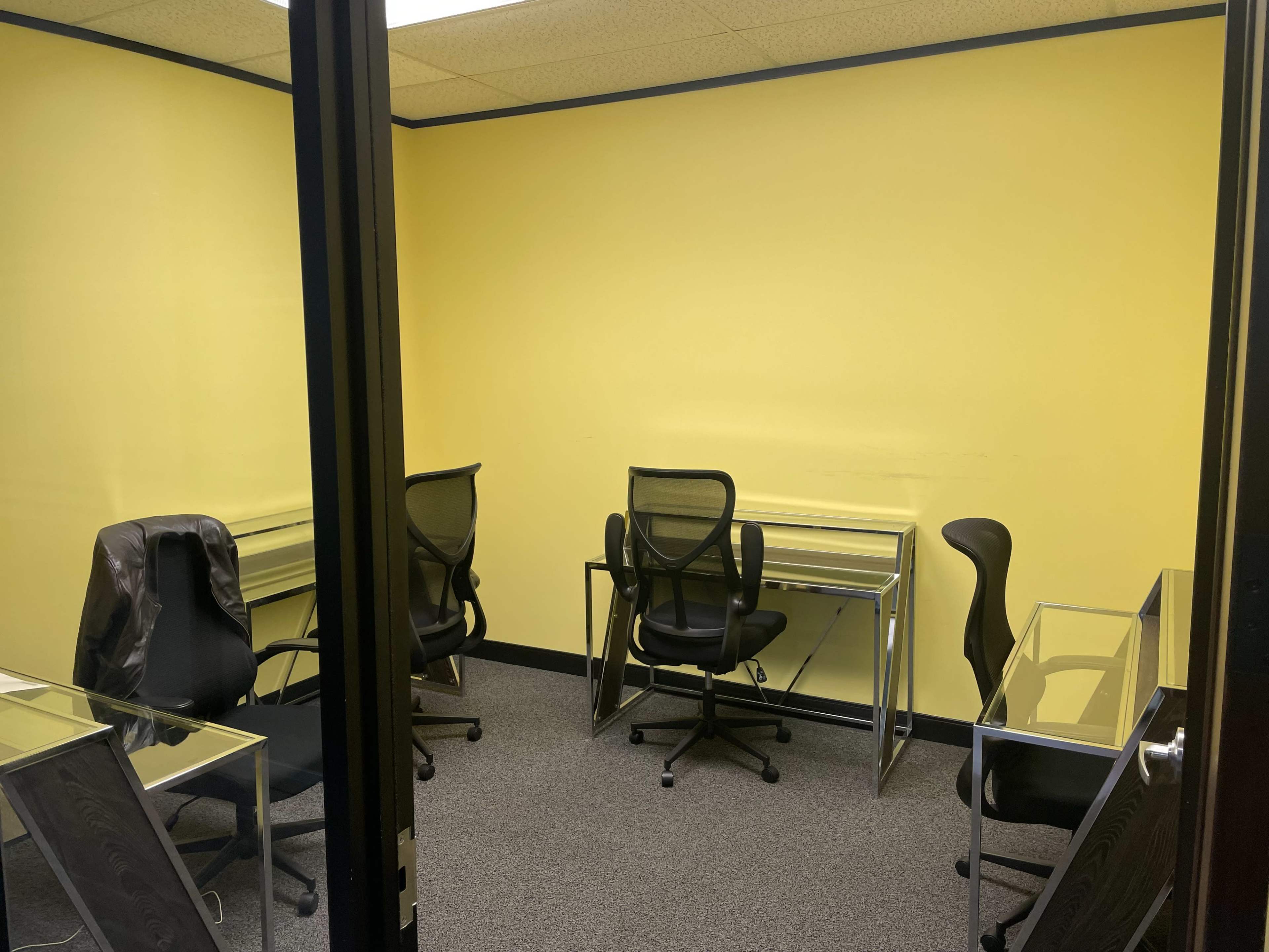 A small office space with four glass-top desks and ergonomic chairs against a bright yellow wall.