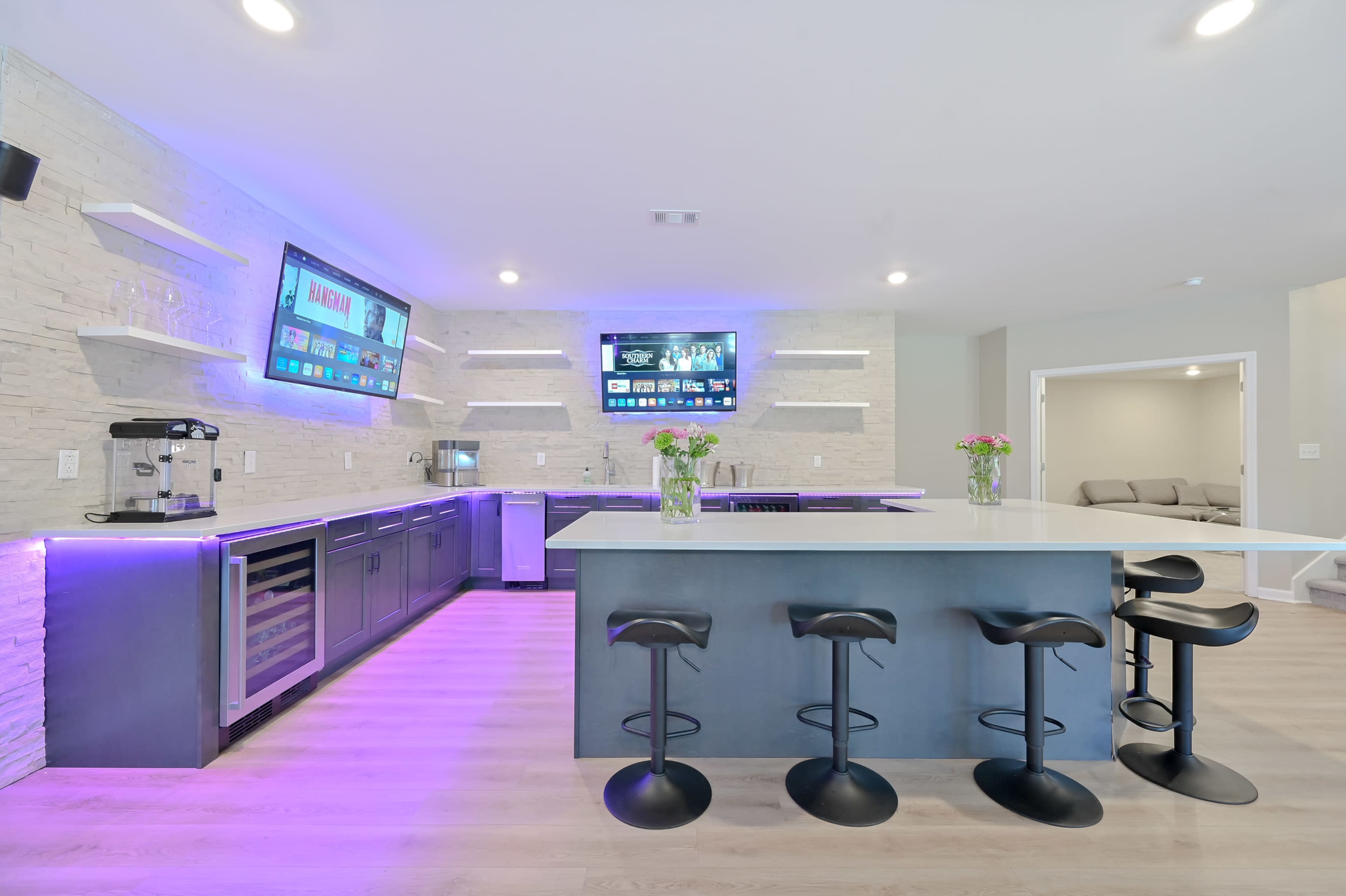 A modern kitchen with a large island, bar seating, and wall-mounted screens displaying information, illuminated with purple lighting.