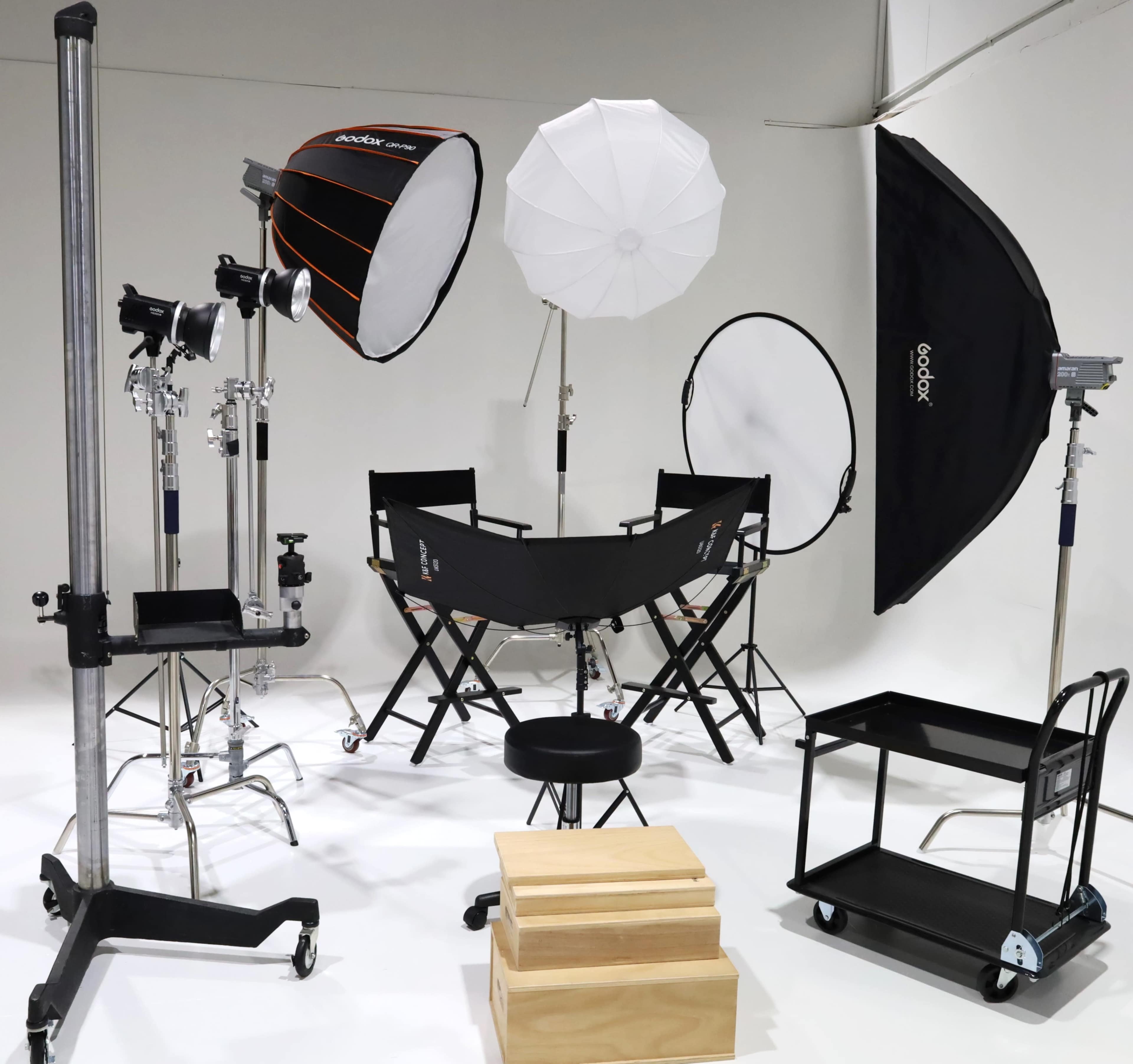 The image shows a photography studio setup with various lighting equipment, reflectors, and support stands arranged around a director's chair and a stool on a white backdrop.