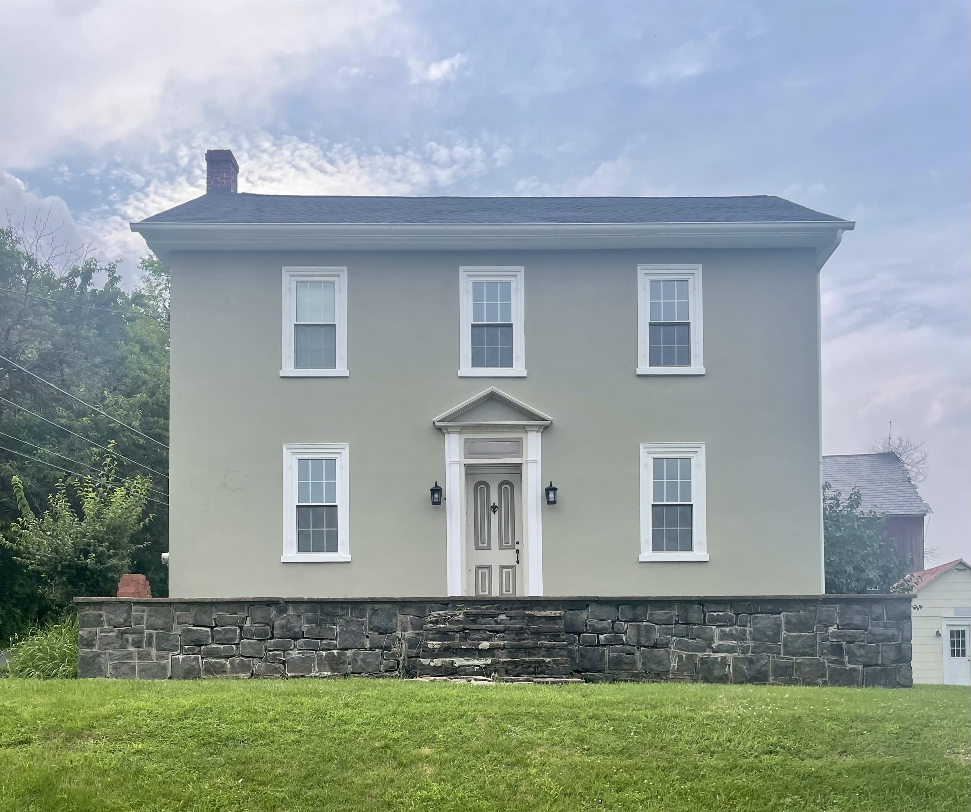 A two-story house with a gray exterior, centered front door, and white trim is situated on a grassy yard with a stone foundation.